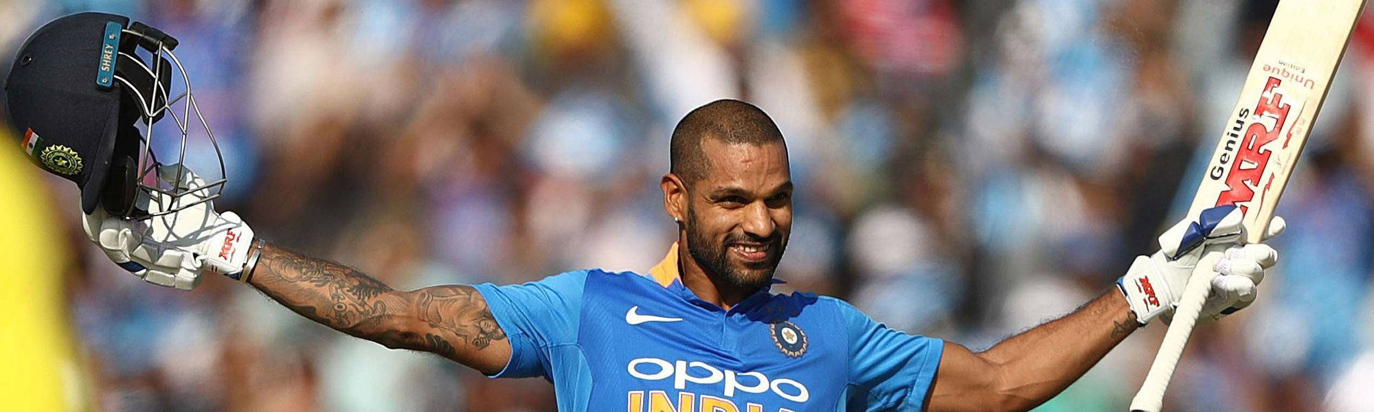 shikhar dhawan 桌布 壁紙 — 印度板球運動員張開雙臂慶祝，手持頭盔和球棒，背景是模糊的體育場人群。