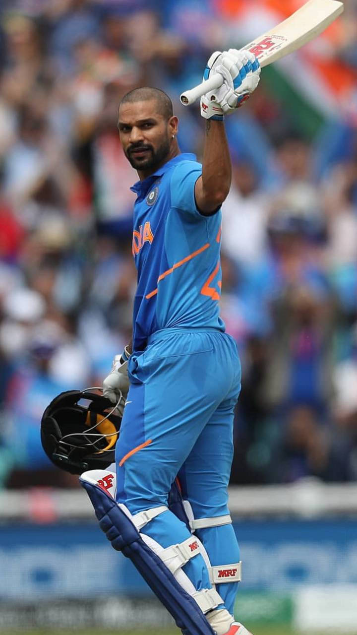 shikhar dhawan 桌布 壁紙 — 印度板球運動員身穿藍色制服，在擠滿觀眾的體育場內高舉球棒，同時手持頭盔和護墊，展現勝利姿態。
