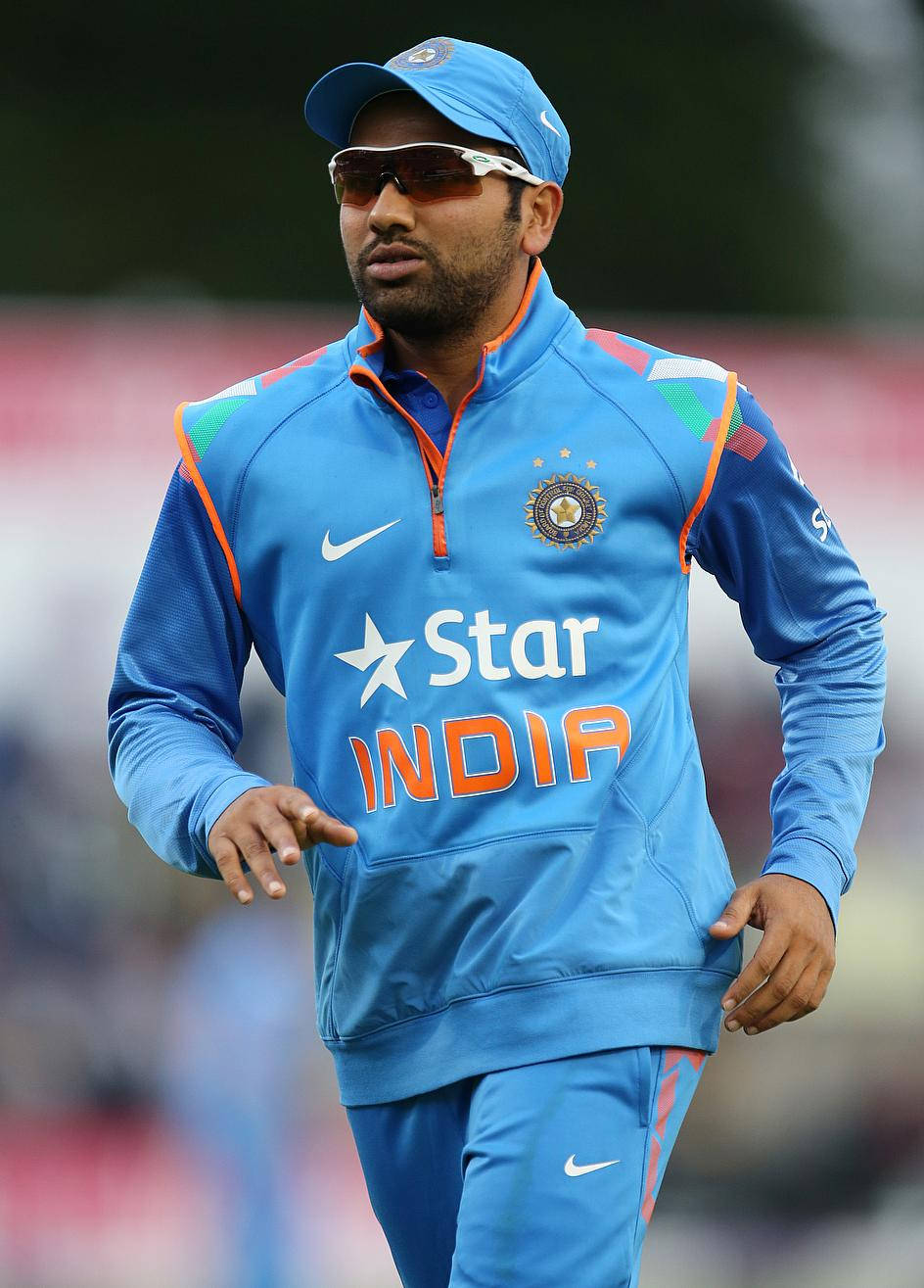 Rohit Sharma 桌布 壁紙 — 印度板球運動員身穿藍色訓練裝備，戴著太陽眼鏡，自信地走在運動場上。