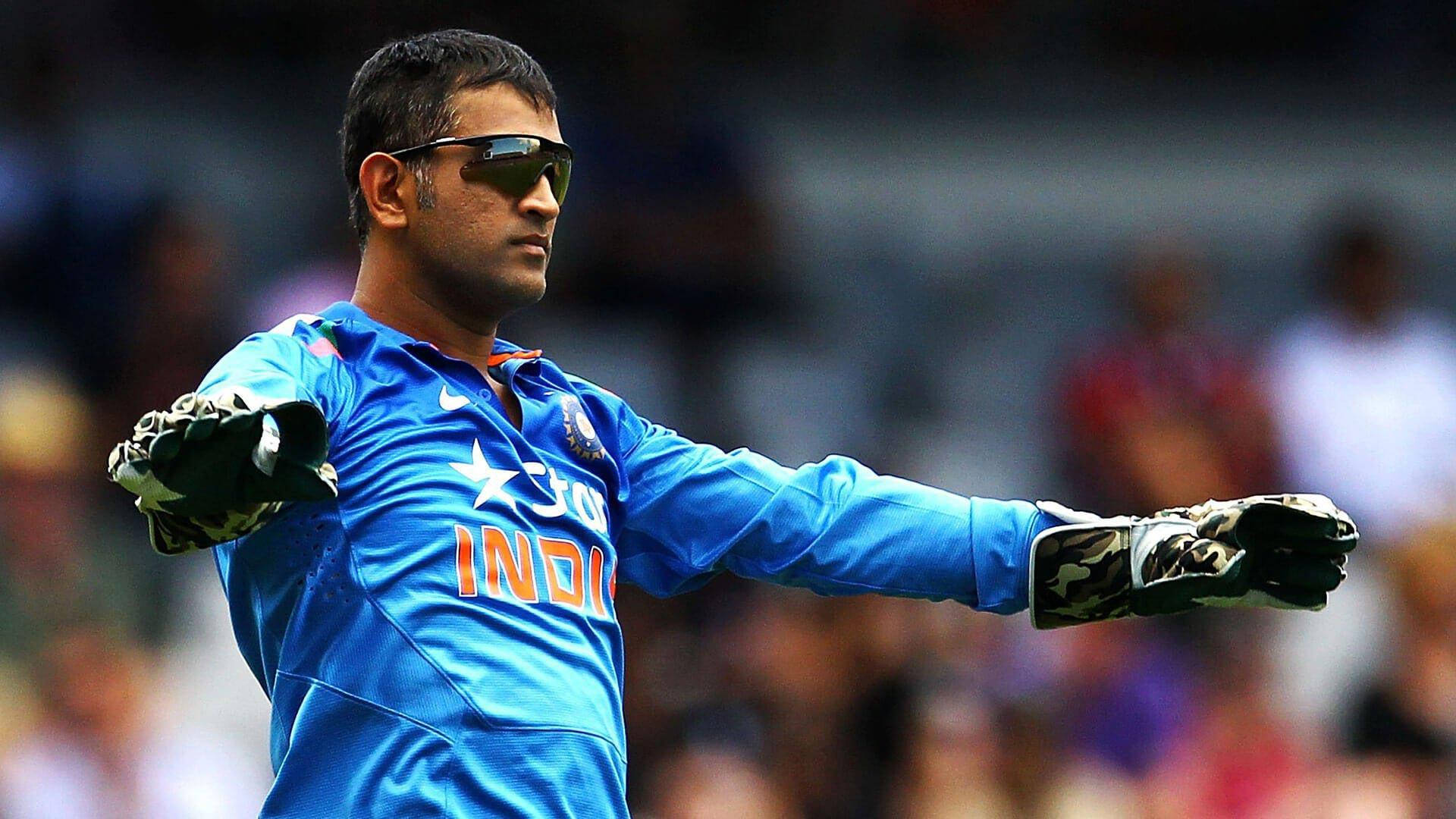 MS Dhoni 桌布 壁紙 — 印度板球隊長 Mahendra Singh Dhoni 身穿藍色球衣和手套，在明亮的體育場燈光下於比賽中做手勢。