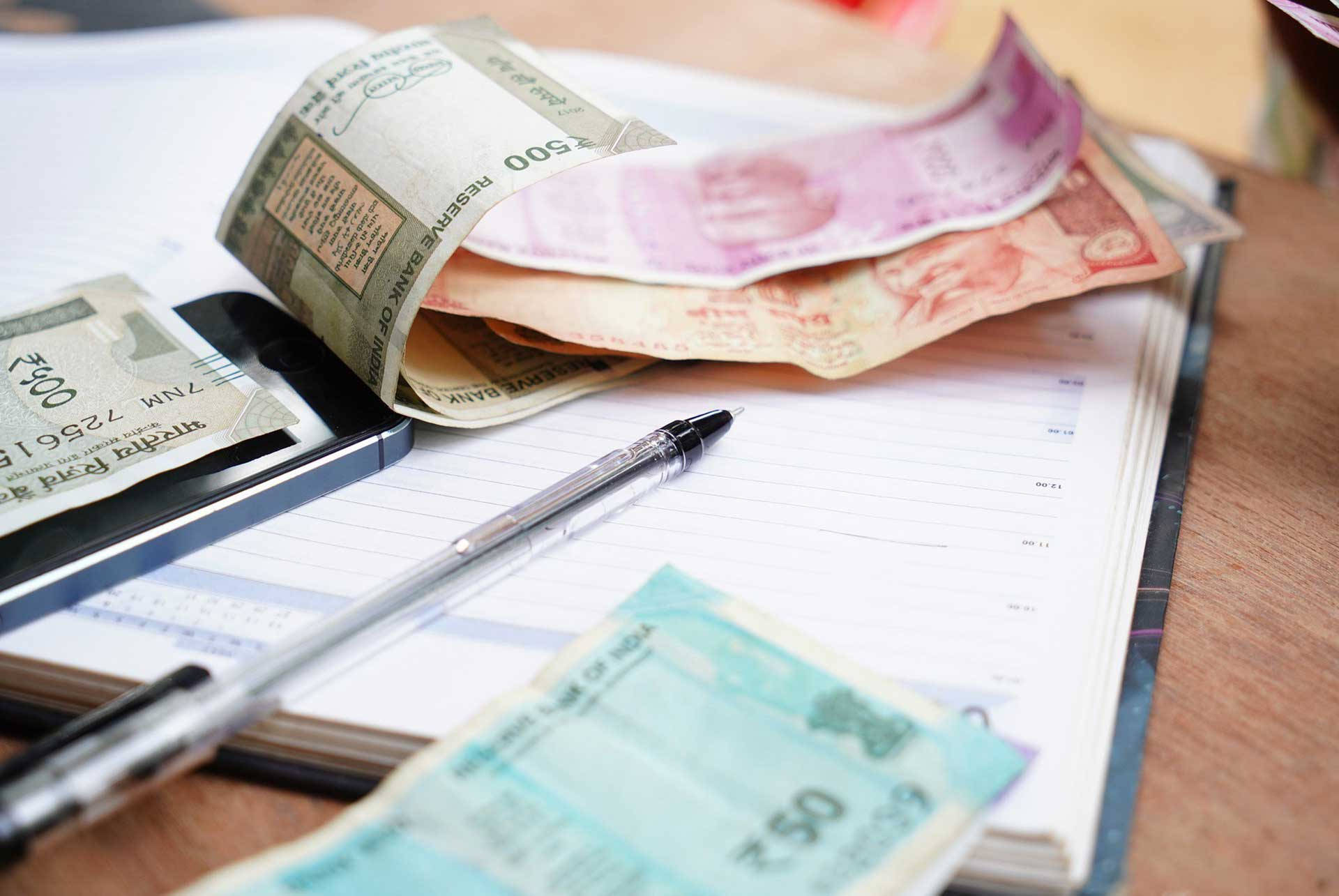 Indian 桌布 壁紙 — 印度盧比鈔票散落在木桌上的分類帳和智慧型手機上，象徵財務規劃或預算編製。