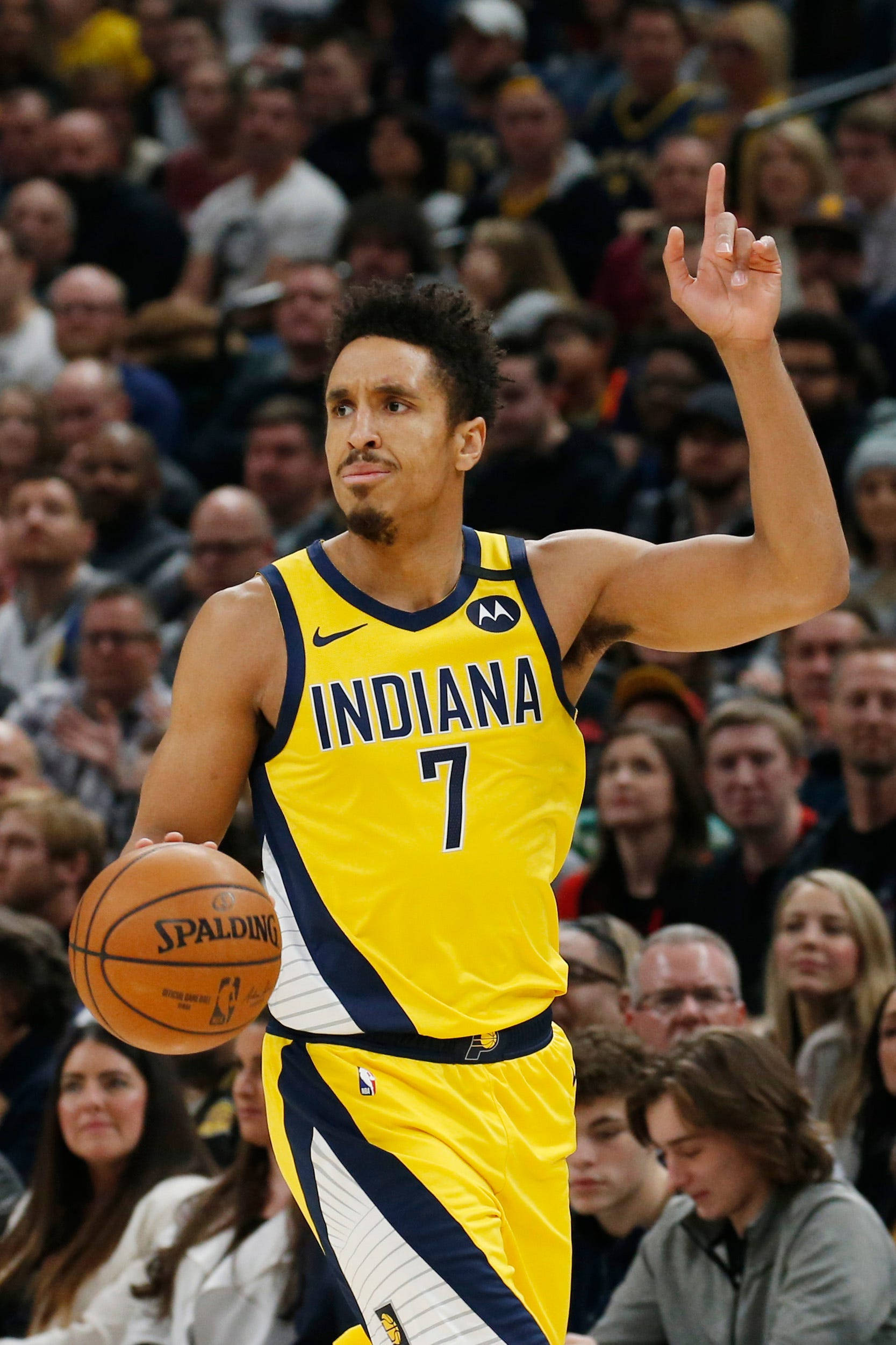 malcolm brogdon 桌布 壁紙 — 印第安納溜馬隊7號球員身穿黃色球衣，在比賽中手持籃球並向上指，背景有觀眾。