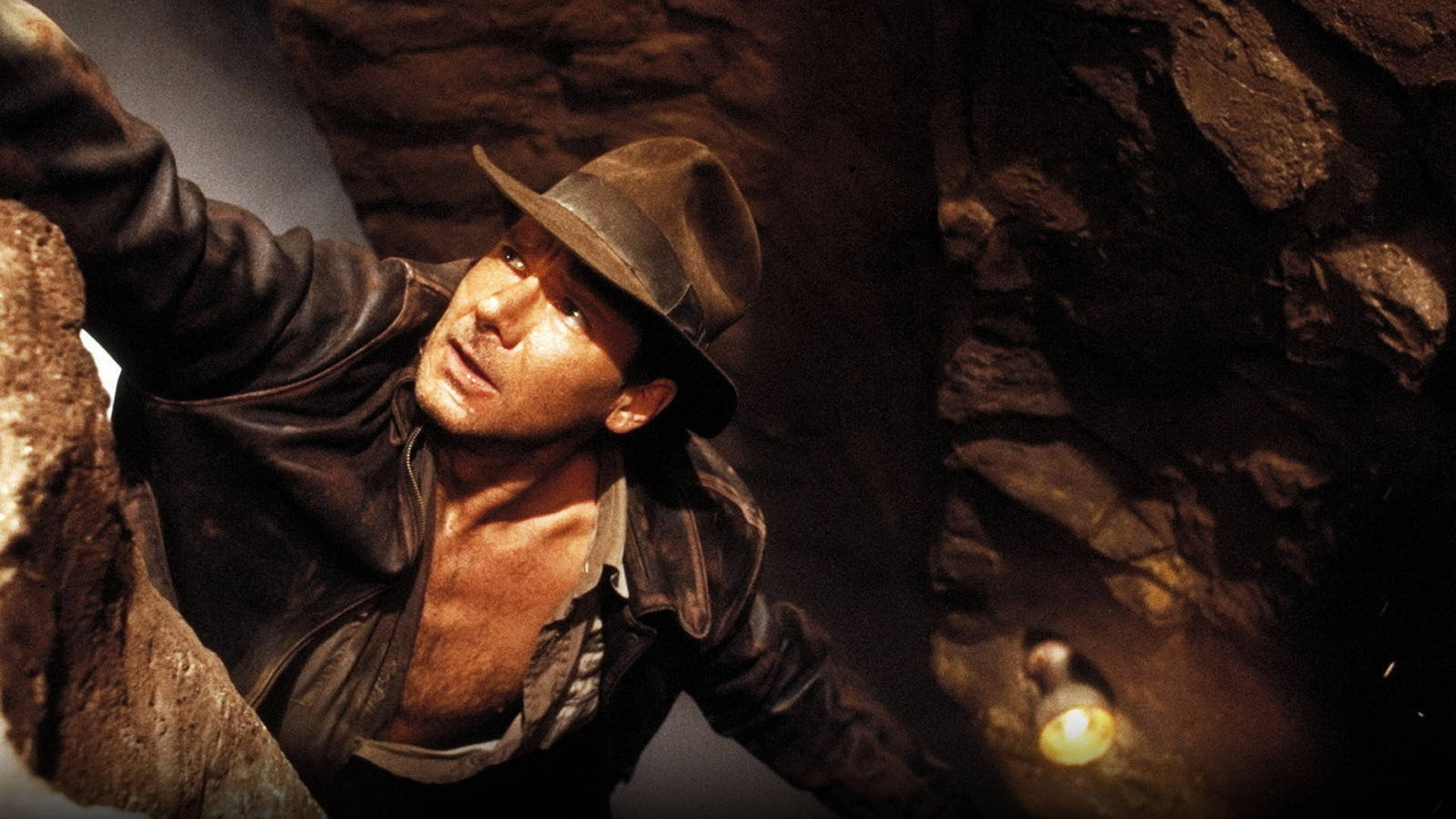 Indiana Jones 桌布 壁紙 — 印第安納瓊斯在昏暗的洞穴中抓住岩石邊緣，他的手電筒照亮了周圍崎嶇的石牆。