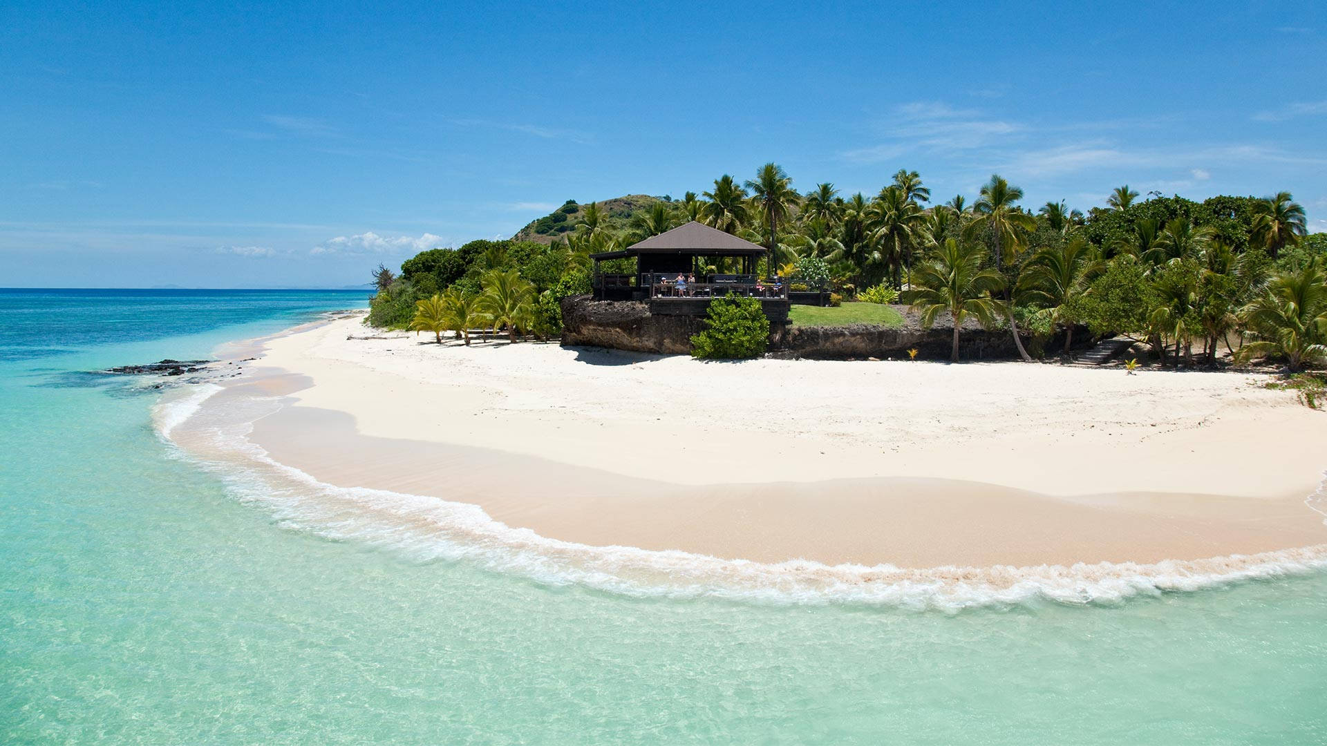 Fiji 桌布 壁紙 — 原始熱帶海灘的鳥瞰圖，擁有碧綠的海水、溫柔的海浪、白色的沙灘和一個坐落在茂密棕櫚樹中的茅草亭。