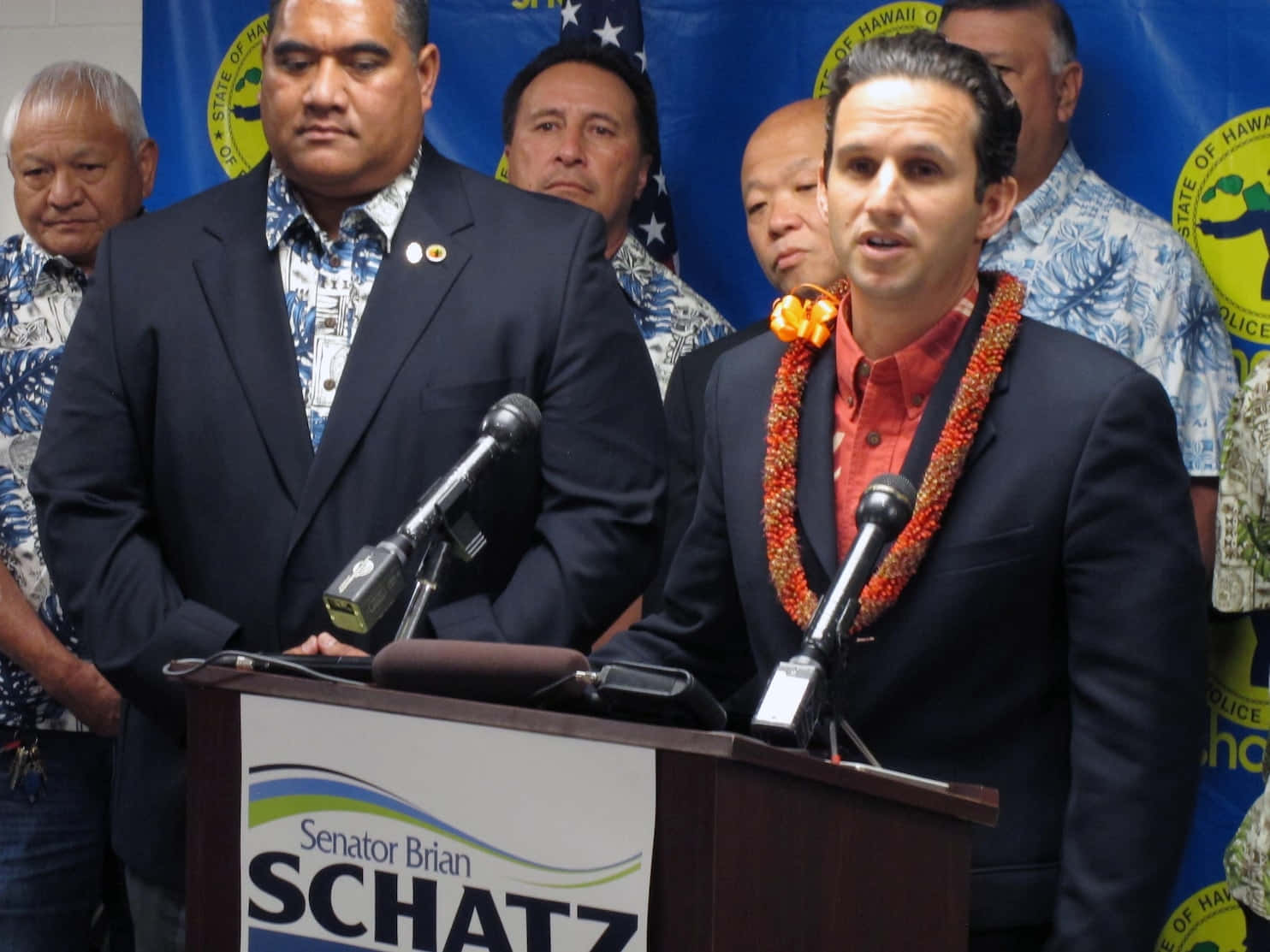 Celebrity 桌布 壁紙 — 參議員 Brian Schatz 在記者會上發言，戴著花環，站在顯示他名字的講台上。