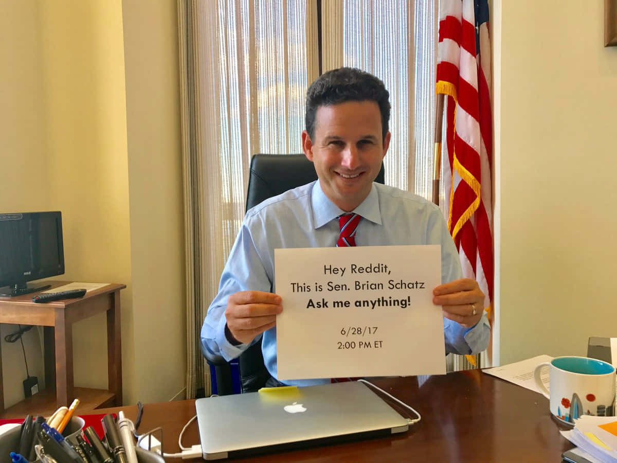Famous 桌布 壁紙 — 參議員Brian Schatz微笑著，在他的辦公室裡拿著一個邀請Reddit問題的牌子，身後有美國國旗。