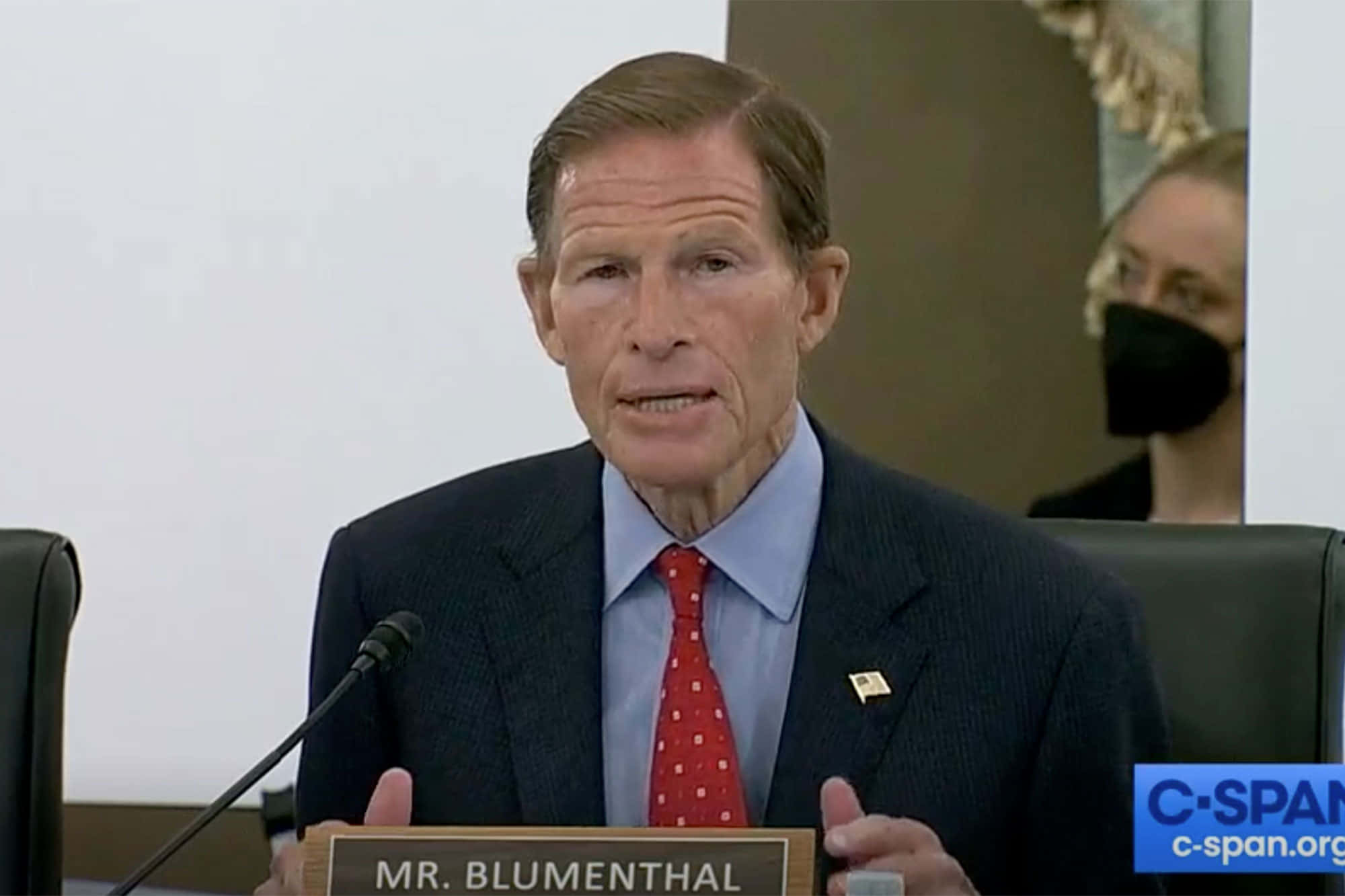 American 桌布 壁紙 — 參議員Richard Blumenthal在正式聽證會上發言，身穿深色西裝、紅色圓點領帶並佩戴名牌。