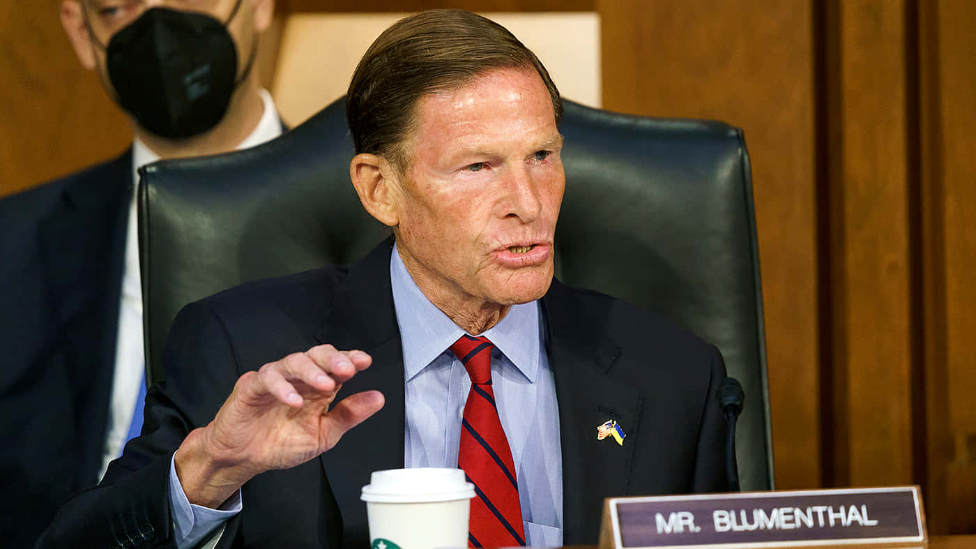 American 桌布 壁紙 — 參議員理查德·布盧門撒爾在正式聽證會上誠懇發言，身穿西裝和紅色領帶，旁邊放著一杯星巴克。