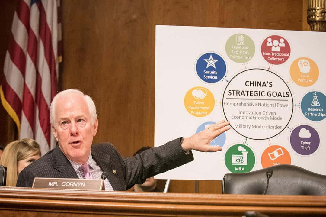 美國 桌布 壁紙 — 參議員約翰·科寧在正式聽證會上指向一張名為「中國的戰略目標」的圖表，背景有美國國旗。