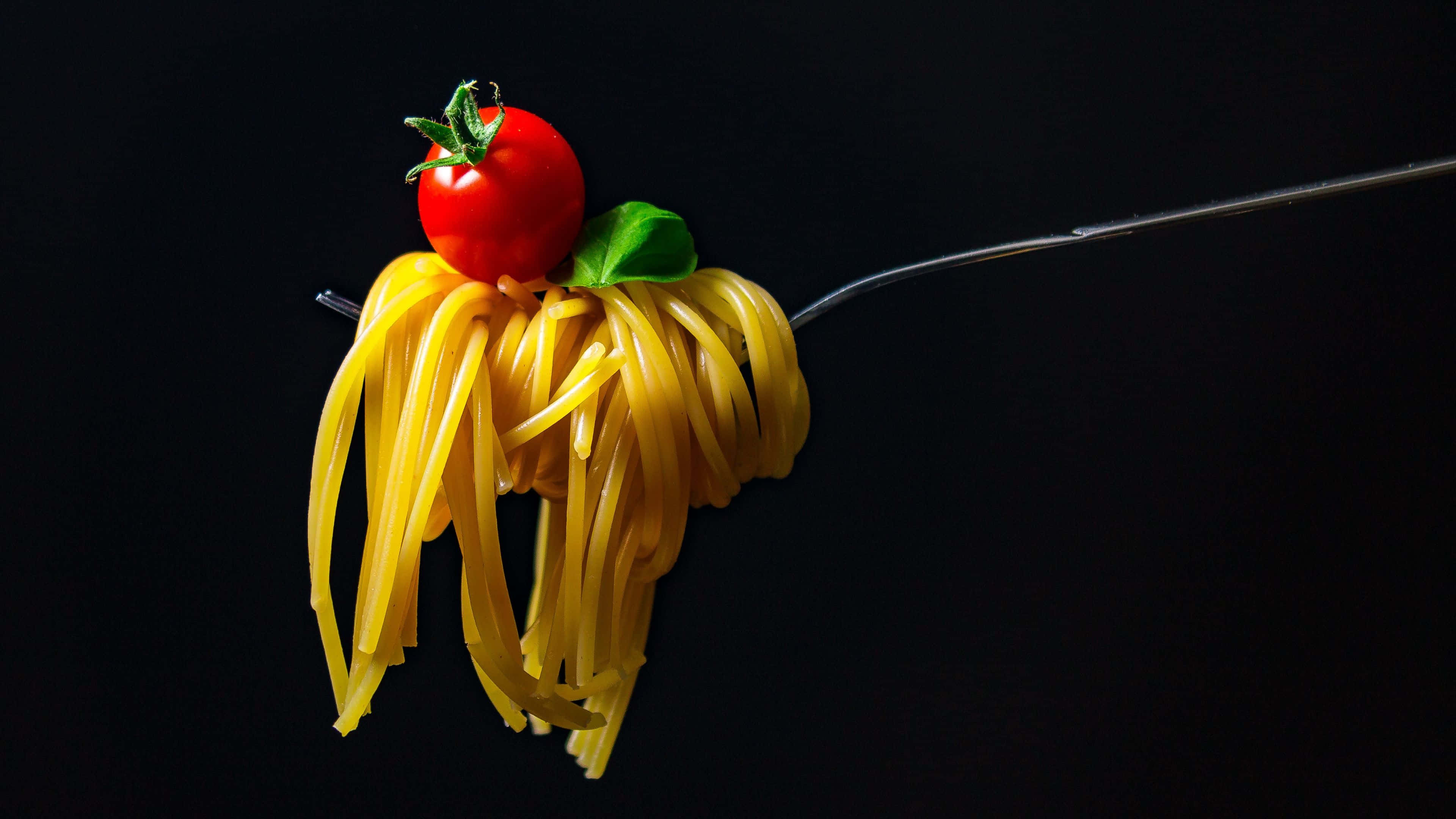 Noodles 桌布 壁紙 — 叉子叉住金黃色的義大利麵，上面放著鮮紅色的櫻桃番茄和綠色的羅勒葉，背景是純黑色。