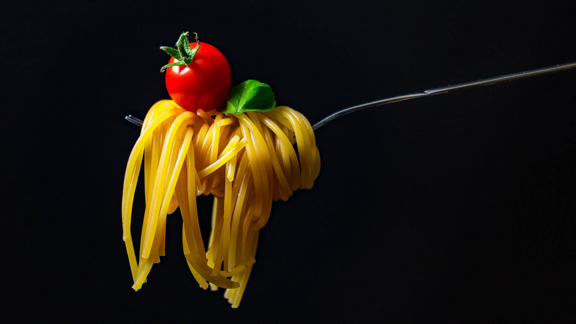 Pasta 桌布 壁紙 — 叉子叉著金黃色的義大利麵，上面放著一顆鮮紅的櫻桃番茄和一片綠色的羅勒葉，背景是純黑色。
