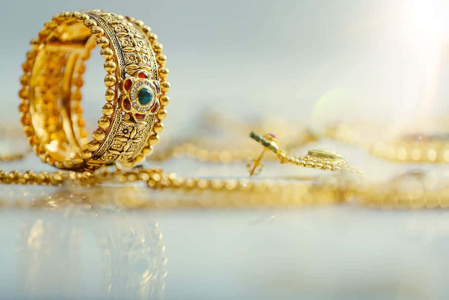 Jewelry Design 桌布 壁紙 — 反射表面上帶有複雜珠飾和中央藍色寶石的華麗黃金首飾特寫。