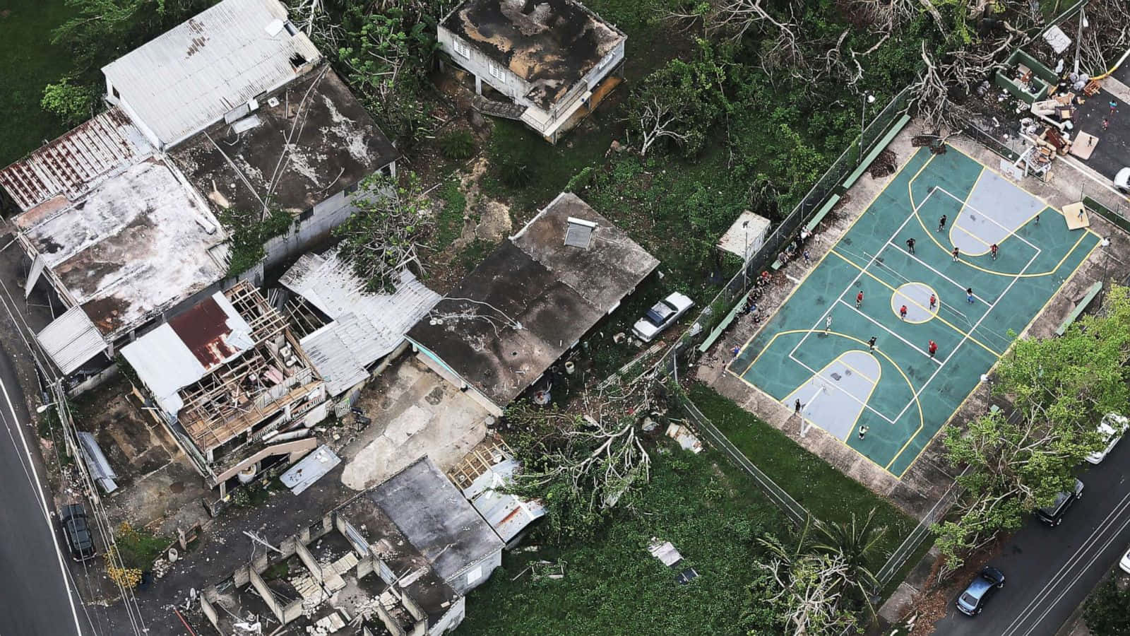 Hurricane 桌布 壁紙 — 受損房屋和人們在充滿活力的綠色籃球場上玩耍的風化社區的鳥瞰圖。