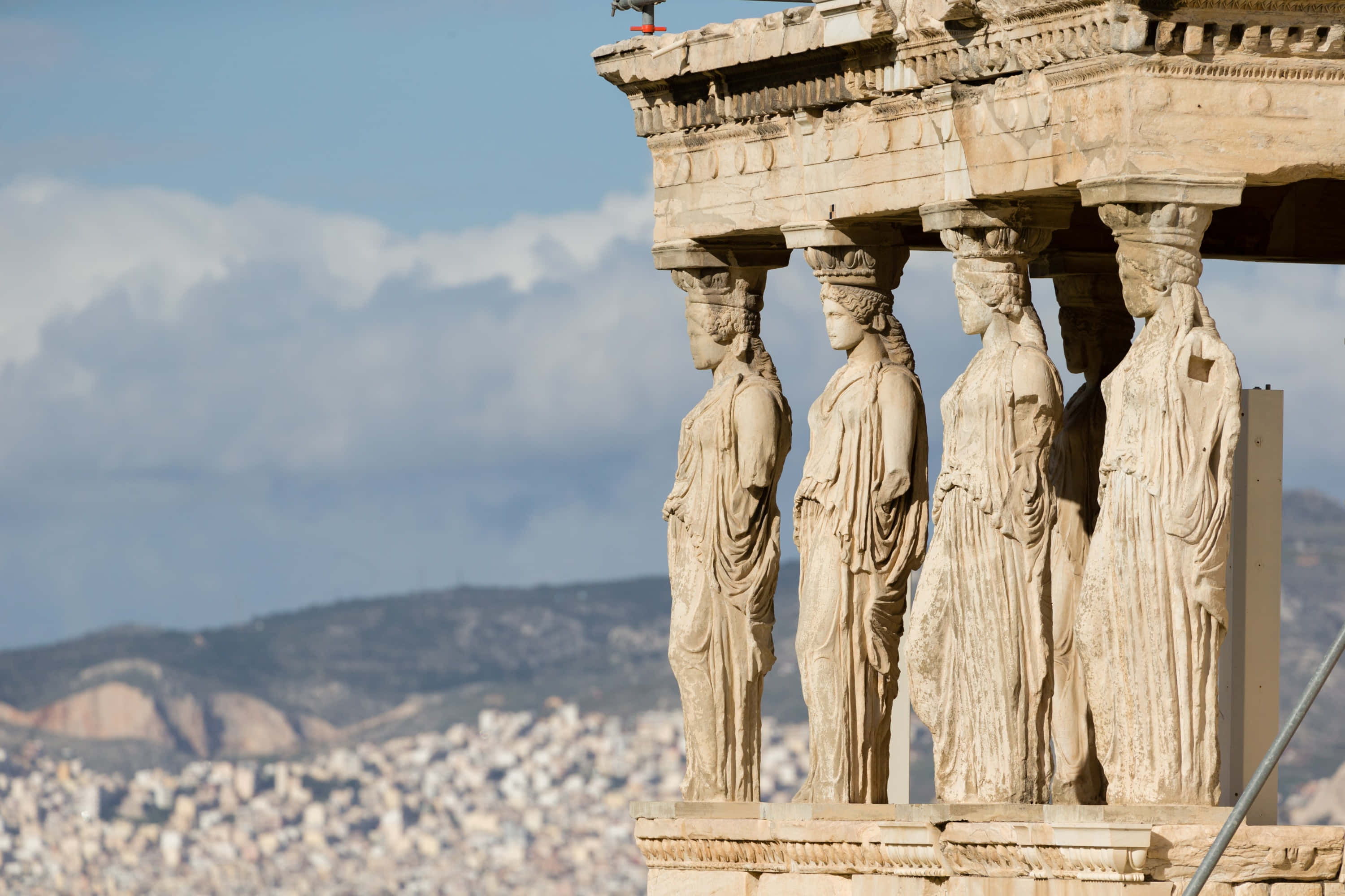 greek 桌布 壁紙 — 古代大理石女像柱支撐著神廟屋頂，背景是雅典城市景觀和遠處的山丘，天空部分多雲。