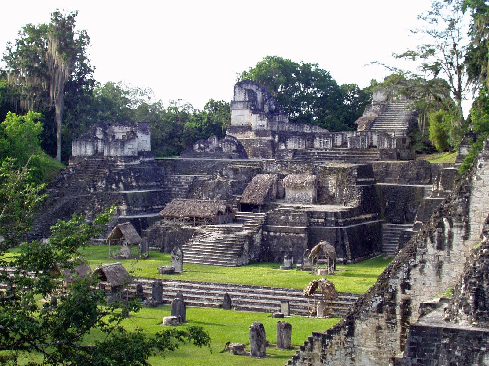 Guatemala 桌布 壁紙 — 古代瑪雅石頭遺跡，階梯金字塔和茅草屋坐落在茂密的綠色叢林中，在明亮的日光下。