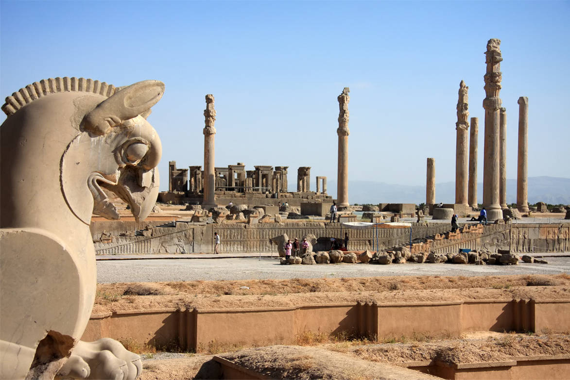 Iran 桌布 壁紙 — 古代石柱和巨大的有翼公牛雕像矗立在陽光普照的考古遺址中，遊客正在探索該地點。