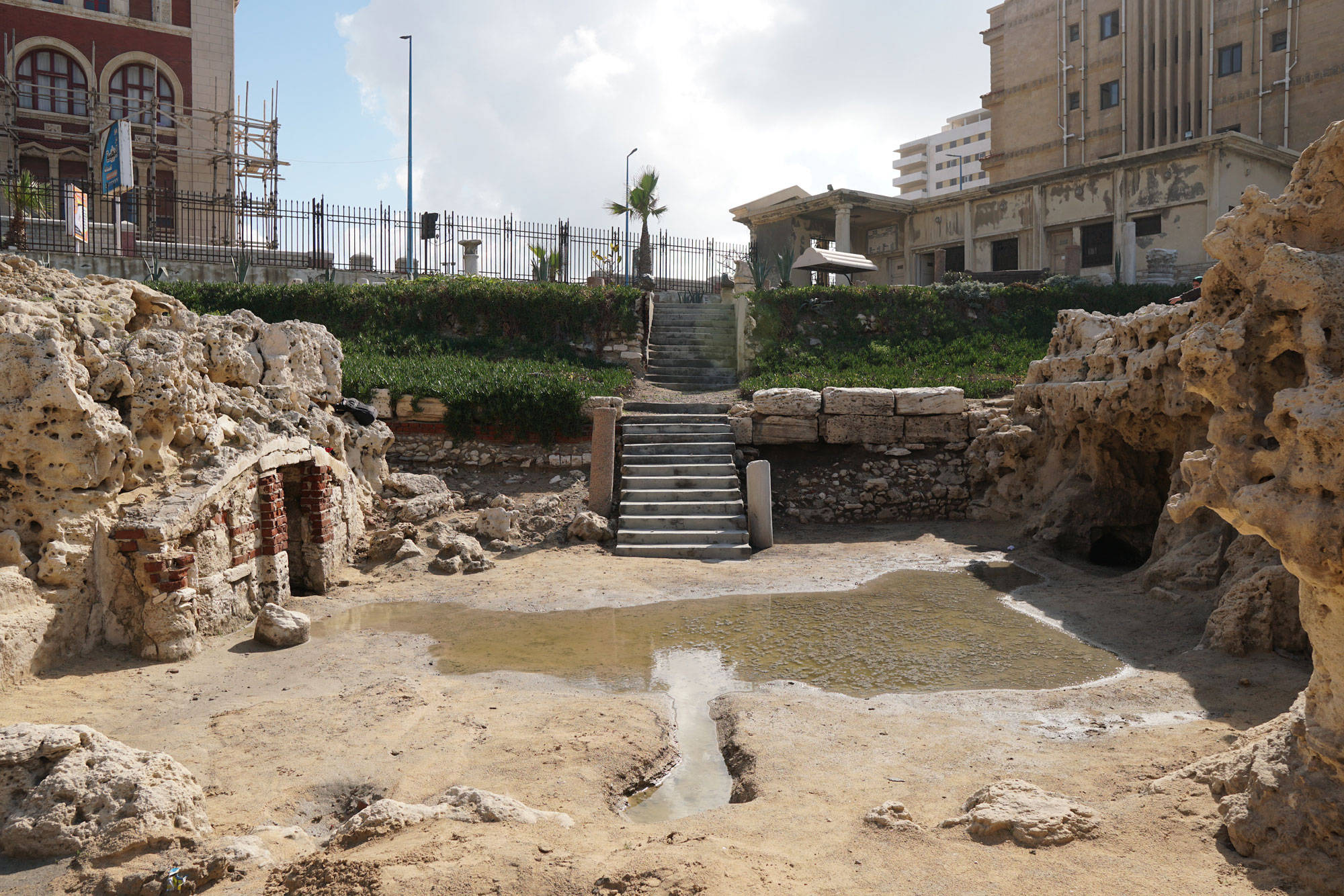 Alexandria 桌布 壁紙 — 古代石頭遺跡，中央有游泳池和樓梯，周圍是現代建築，天空明亮。