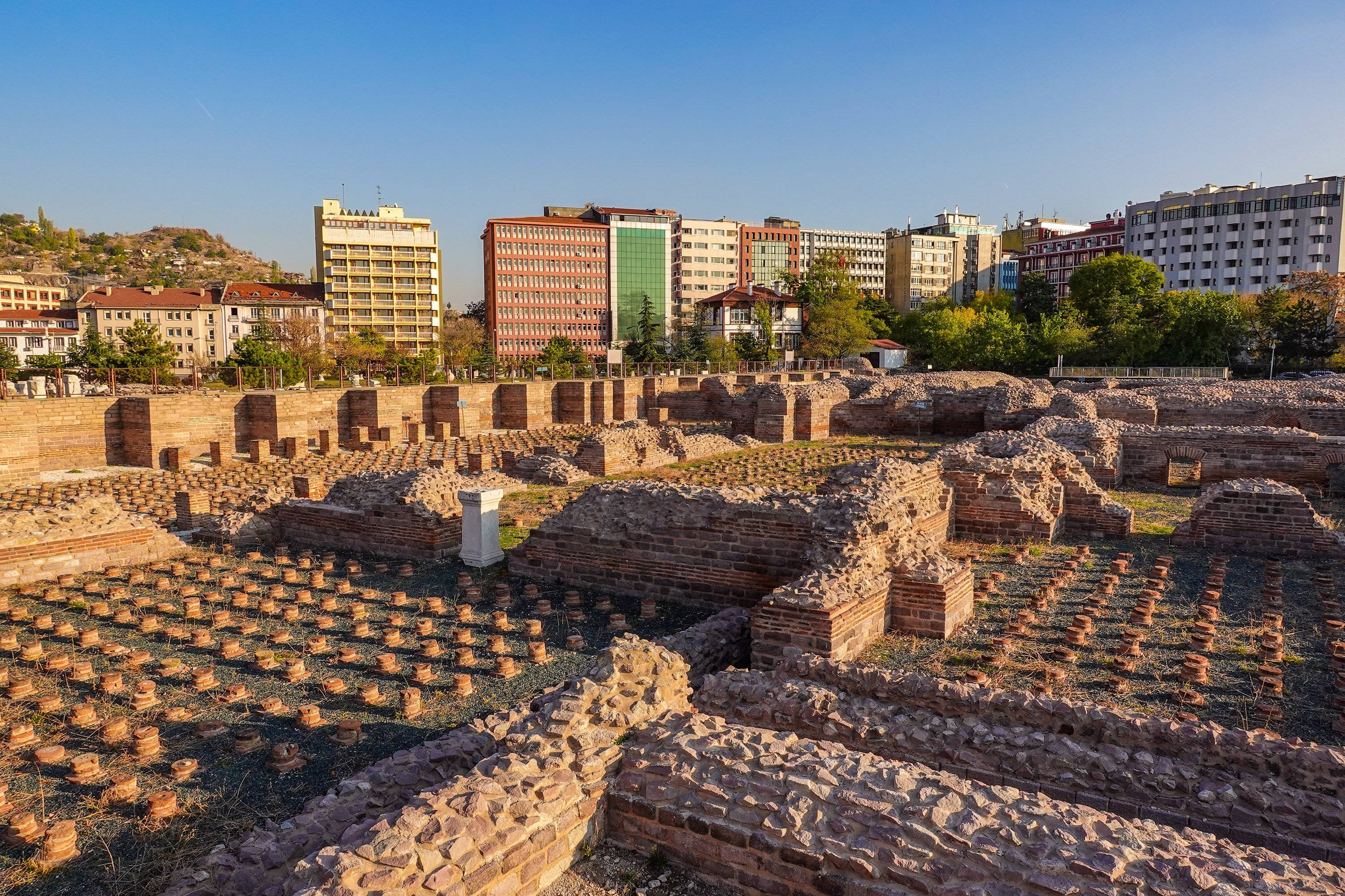 Ankara 桌布 壁紙 — 古代磚砌遺跡，散落著柱子和石頭地基，在晴朗的藍天下，背景是現代建築。