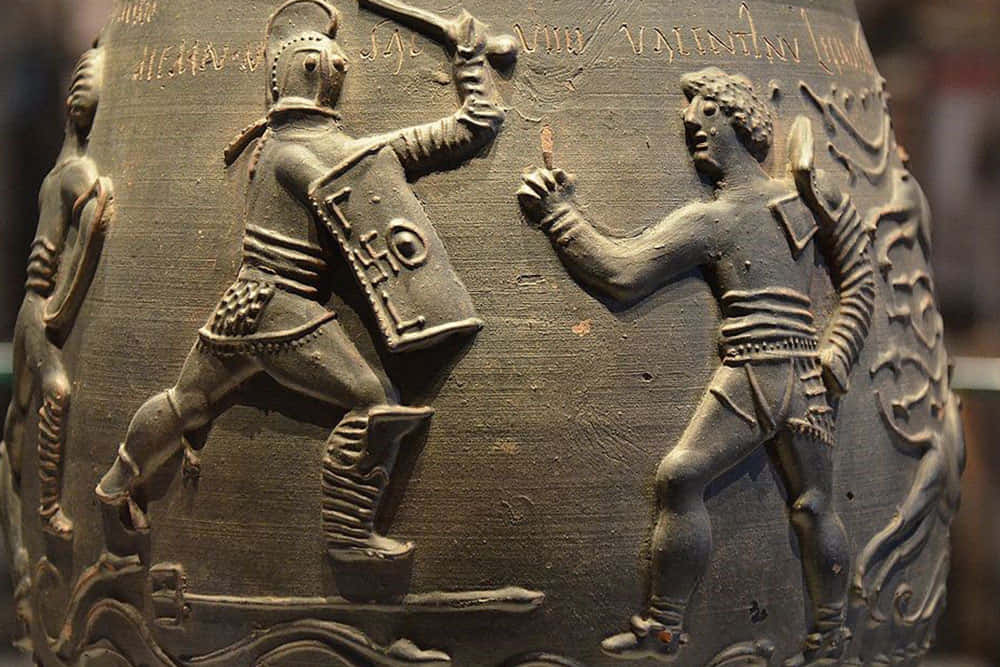 Gladiator 桌布 壁紙 — 古代青銅浮雕特寫，描繪兩名戰士身穿精細盔甲並帶有銘文進行戰鬥。