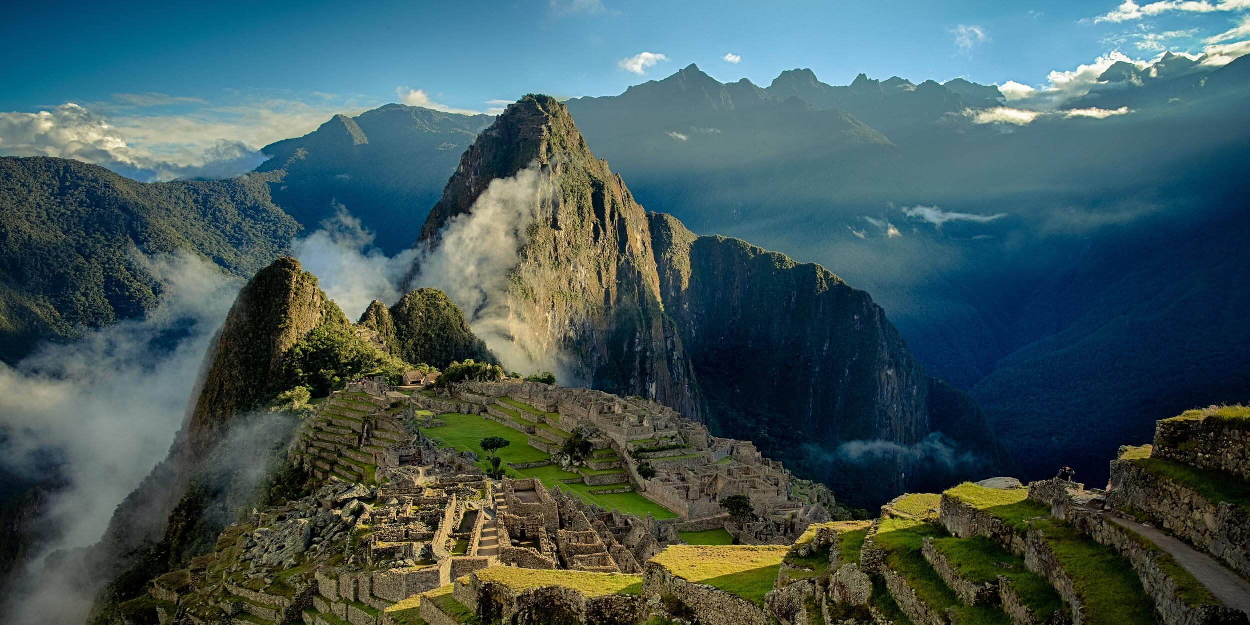 Mountain Landscape 桌布 壁紙 — 古代馬丘比丘遺址依附在霧氣繚繞的山脊上，天空湛藍，雲朵稀疏。