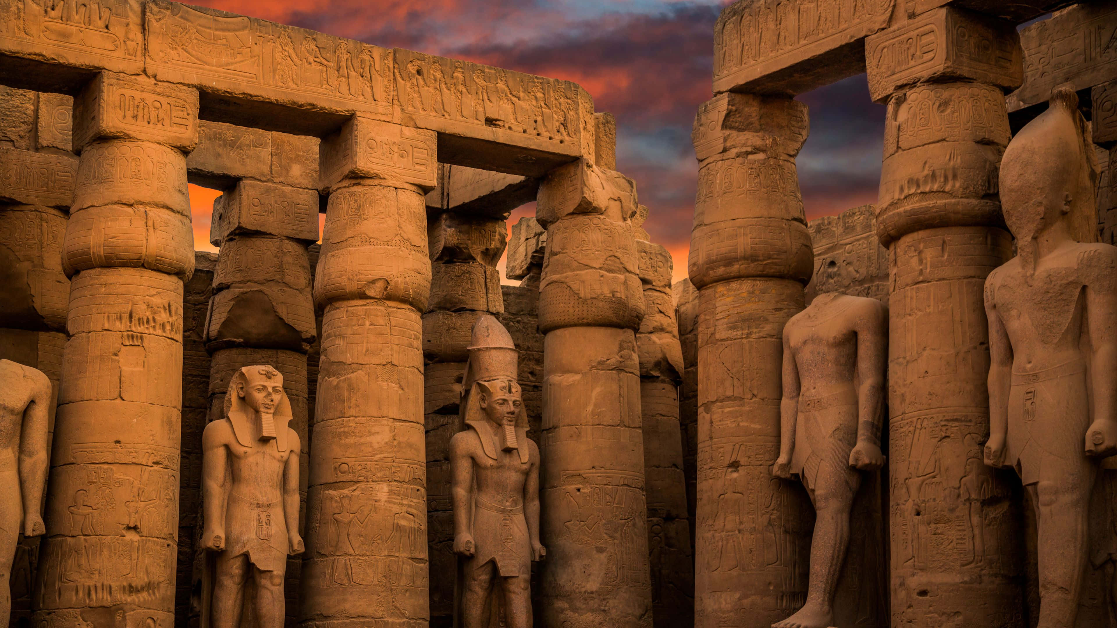 Ancient Egypt 桌布 壁紙 — 古埃及神廟的柱子和雕像沐浴在溫暖的日落光線中，映襯著火熱的橙色和紫色天空。