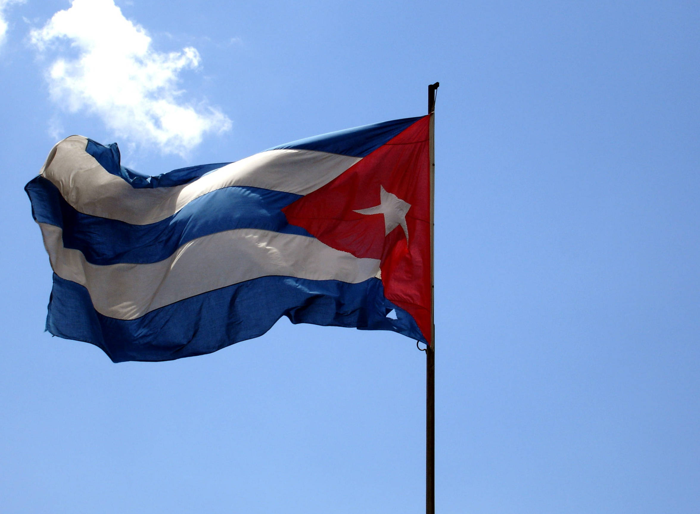 Cuban 桌布 壁紙 — 古巴國旗，帶有紅、白、藍條紋和一顆白色星星，在晴朗的藍天中動態飄揚，天空中有一朵蓬鬆的白雲。