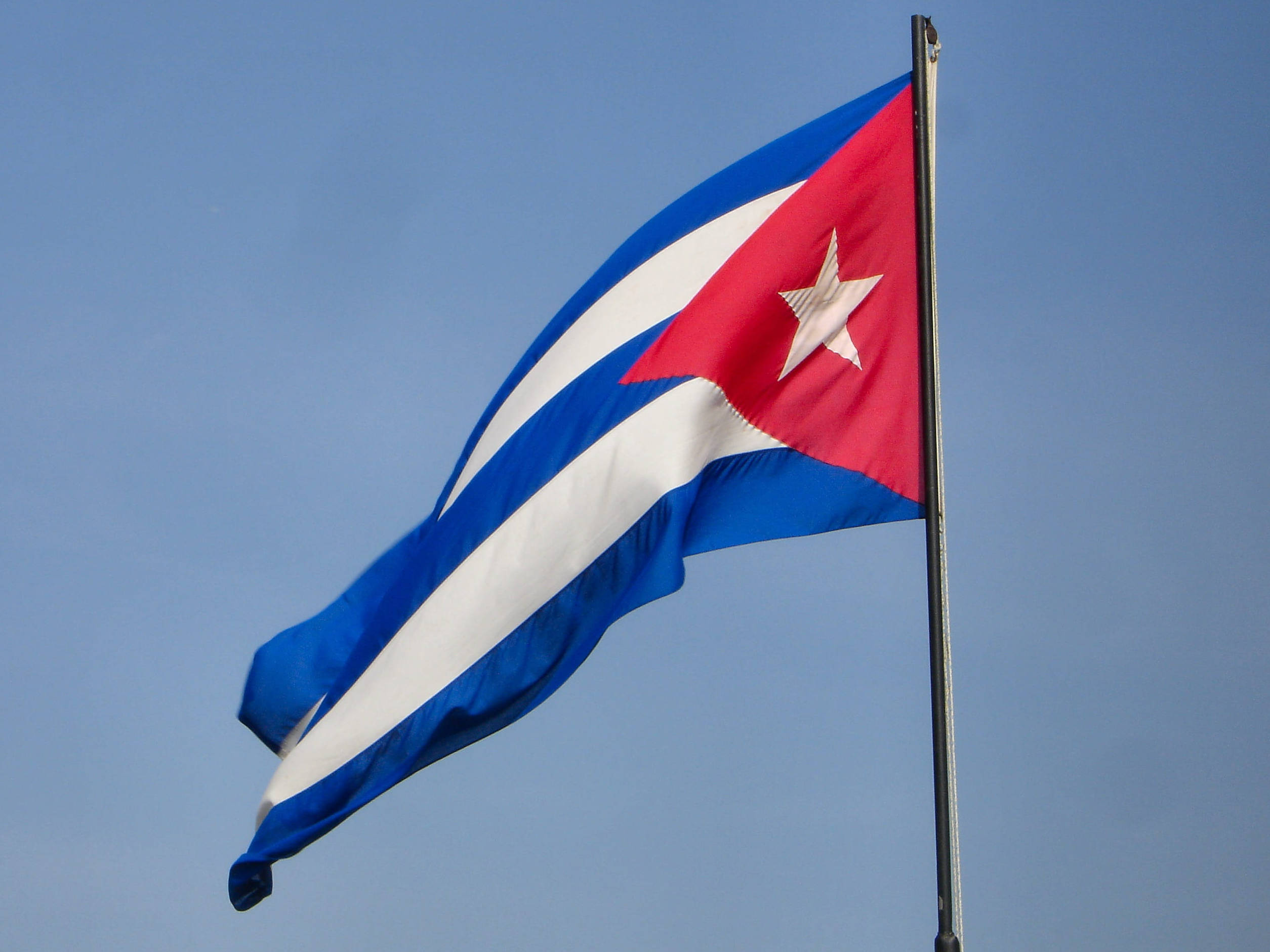 Cuban 桌布 壁紙 — 古巴國旗，有紅、白、藍條紋和一顆白星，在晴朗的藍天下動態飄揚。