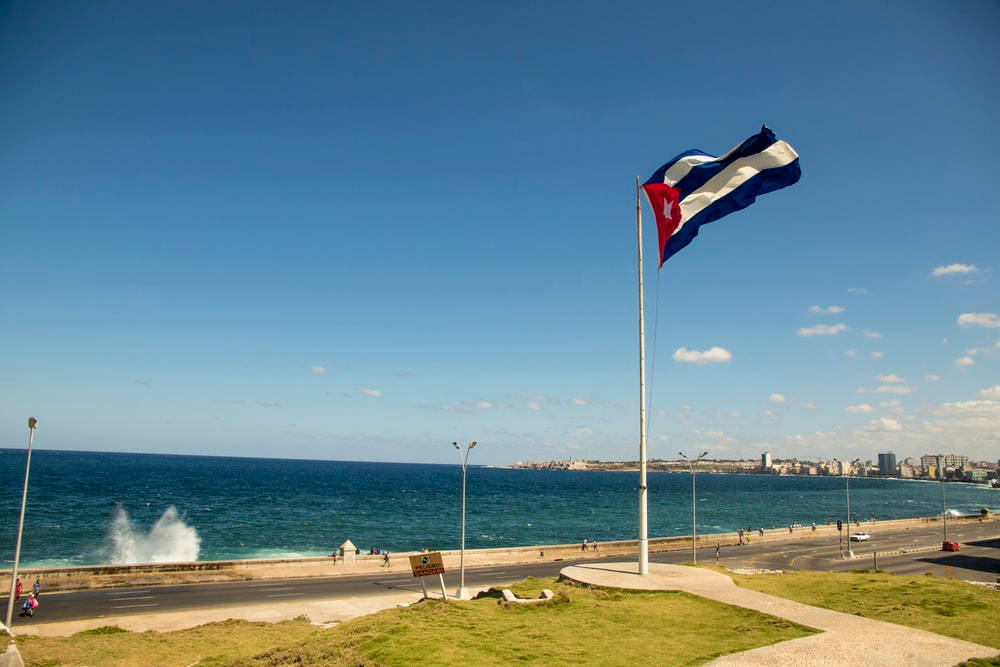 Cuban 桌布 壁紙 — 古巴國旗在旗杆頂端驕傲飄揚，俯瞰著充滿活力的海濱長廊，碧綠的海洋和遠處的城市天際線在清澈的藍天下。