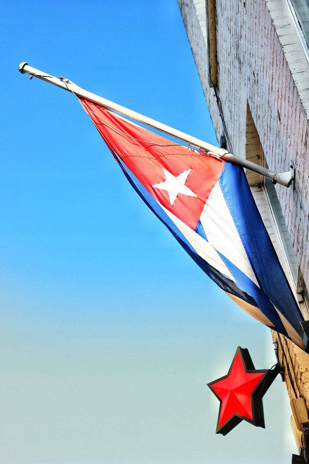 Cuban 桌布 壁紙 — 古巴國旗在晴朗的藍天下，在一棟帶有紅色星星標誌的磚砌建築旁大膽飄揚。