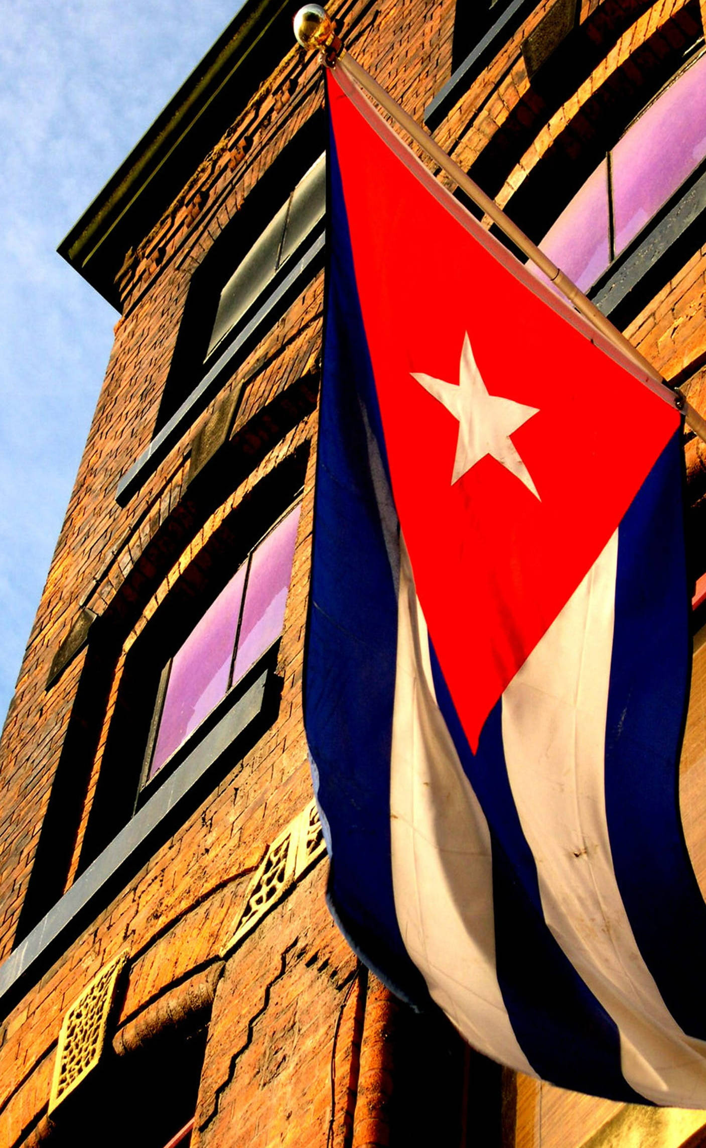 Cuban 桌布 壁紙 — 古巴國旗在晴朗藍天和溫暖陽光下，在磚砌建築前大膽飄揚。