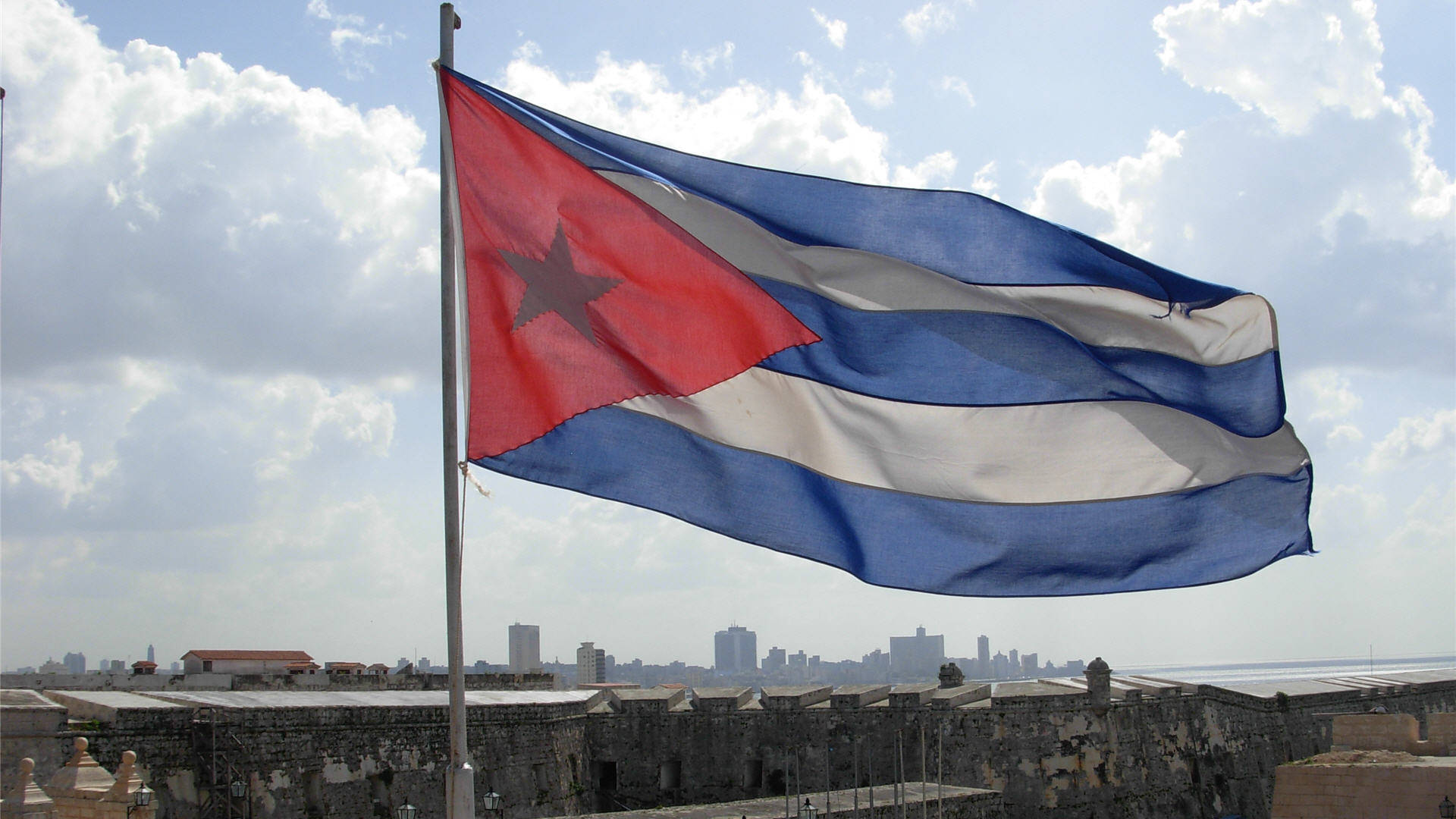 Cuban 桌布 壁紙 — 古巴國旗在歷史悠久的石堡壘頂部自豪地飄揚，城市天際線和海洋在部分多雲的天空下清晰可見。