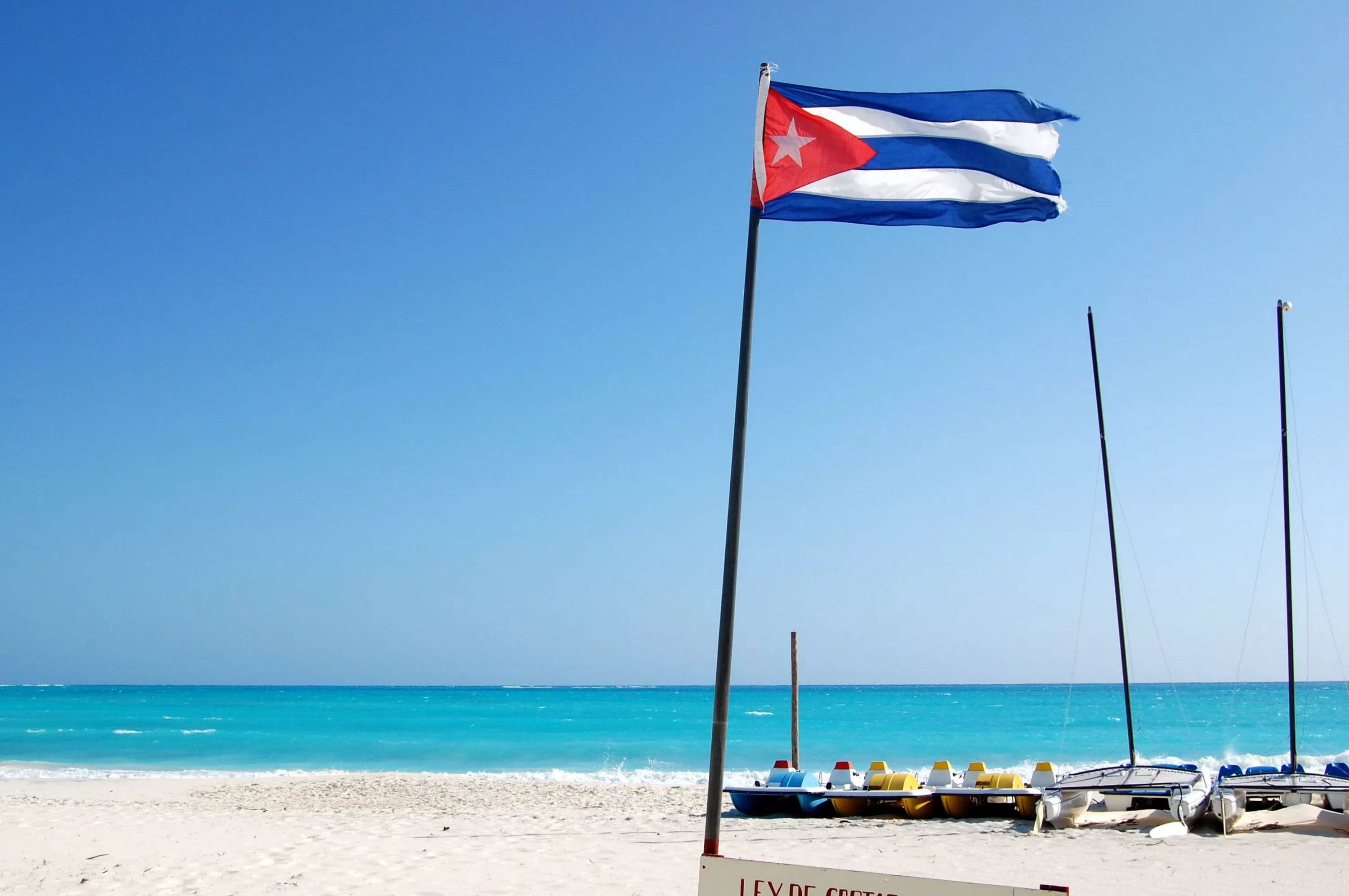Cuba 桌布 壁紙 — 古巴國旗在陽光明媚的海灘上自豪地飄揚，前景是碧綠的海洋和划艇。