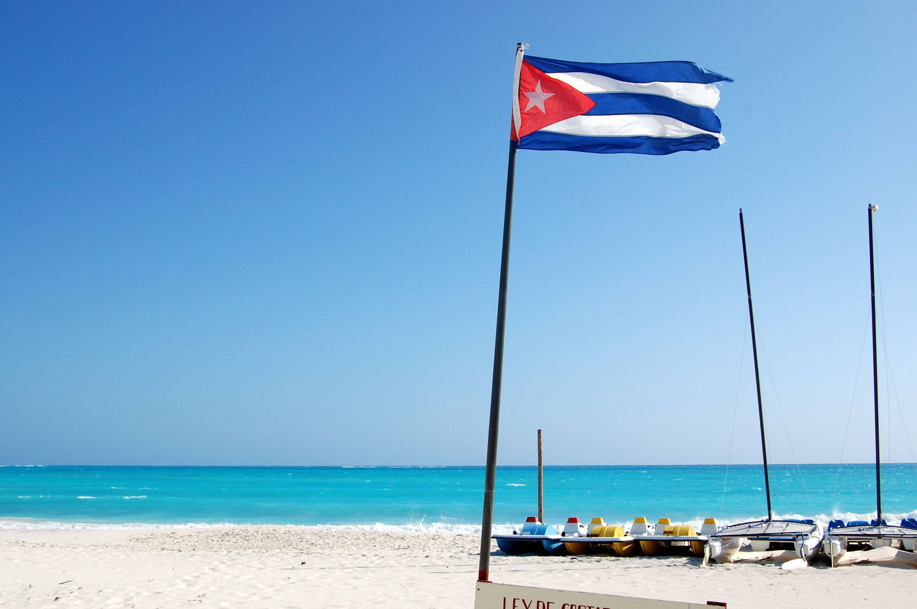 Cuban 桌布 壁紙 — 古巴國旗在陽光明媚的海灘上自豪地飄揚，附近有碧綠的海水和停泊的划艇。