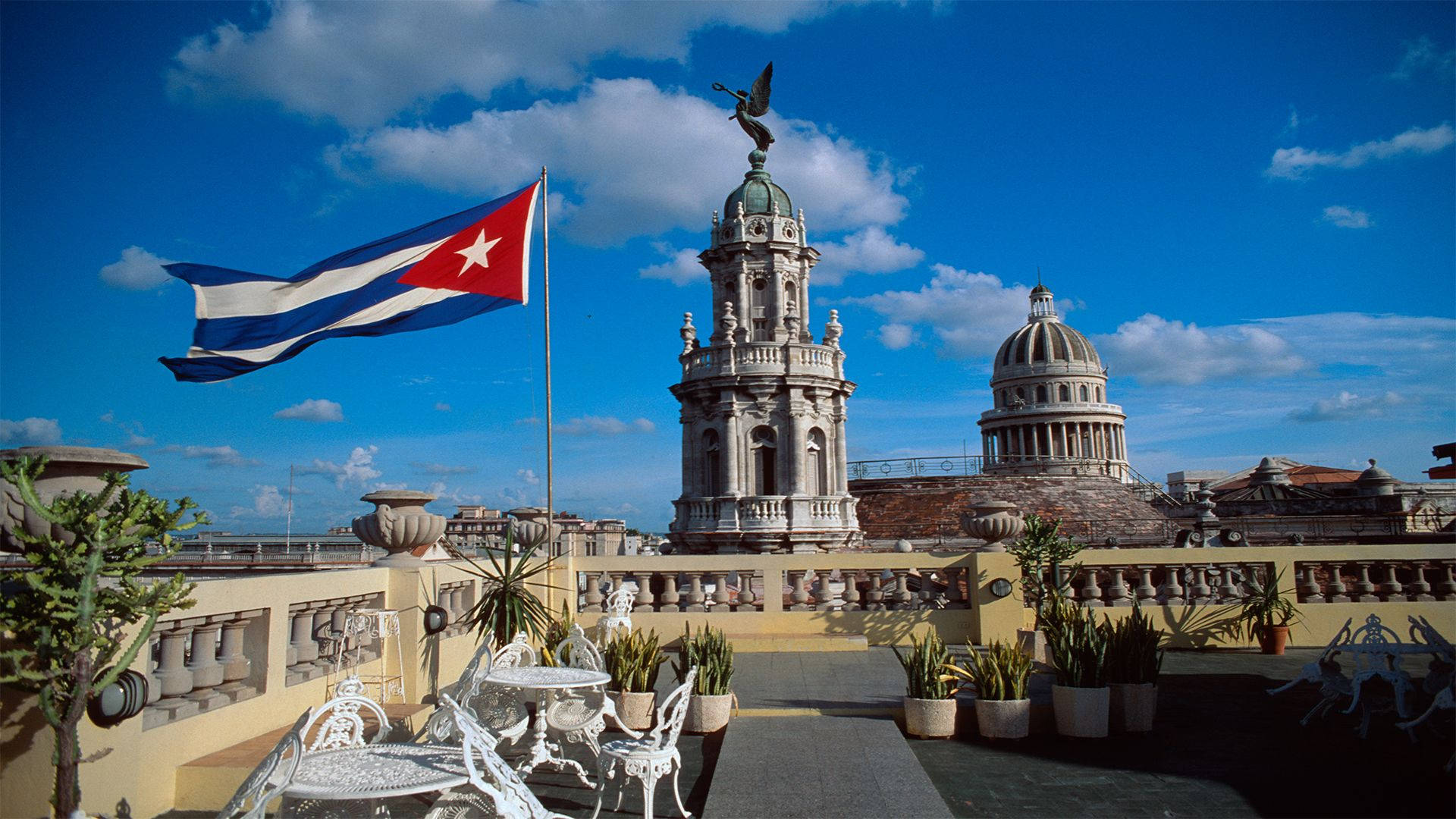 Cuba 桌布 壁紙 — 古巴國旗在陽光普照的屋頂露臺上自豪地飄揚，俯瞰著歷史悠久的圓頂建築，背景是充滿活力的藍天。