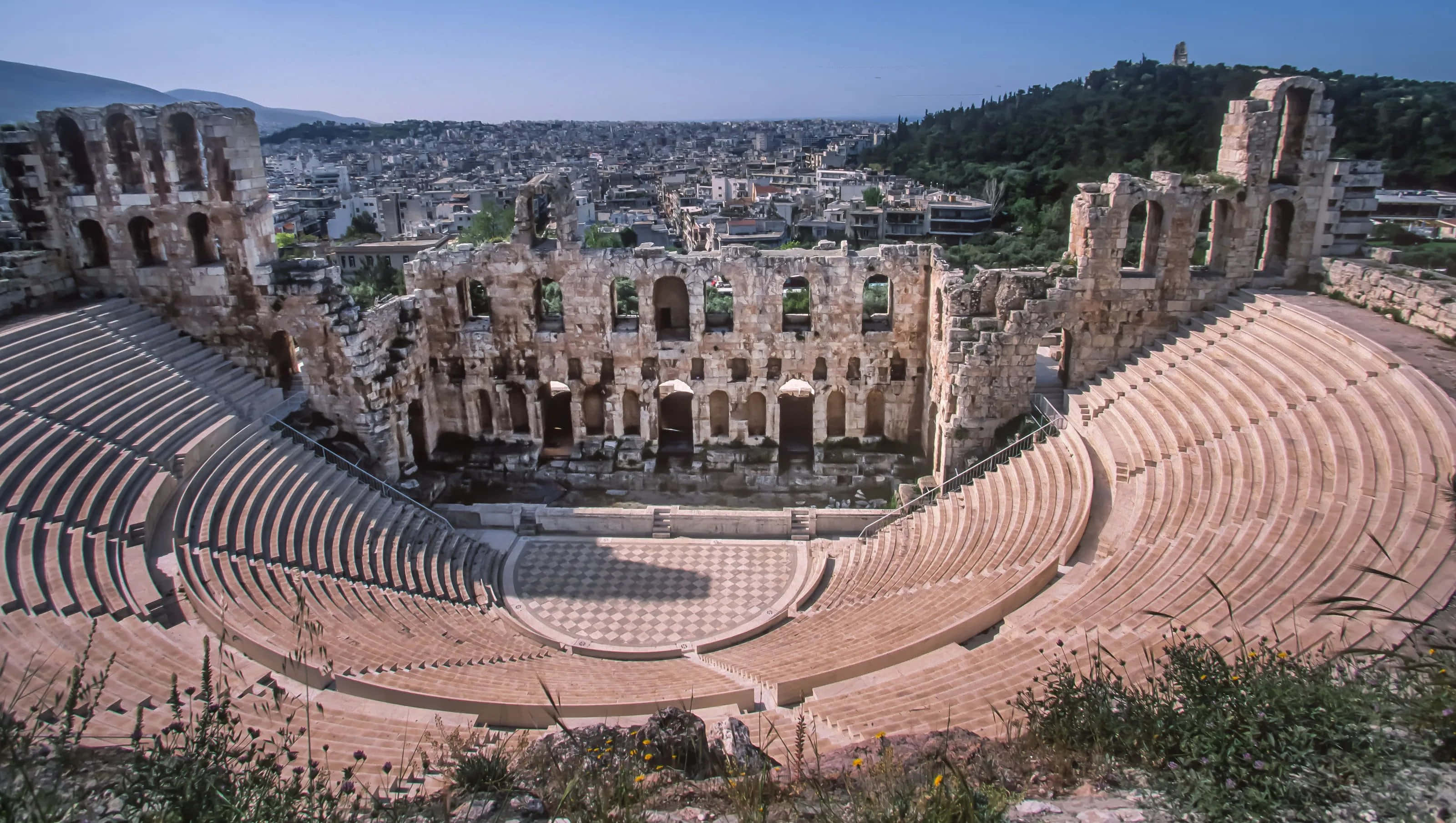 greek 桌布 壁紙 — 古希臘劇場遺址，設有階梯式石製座位，俯瞰晴朗藍天下的現代城市景觀。