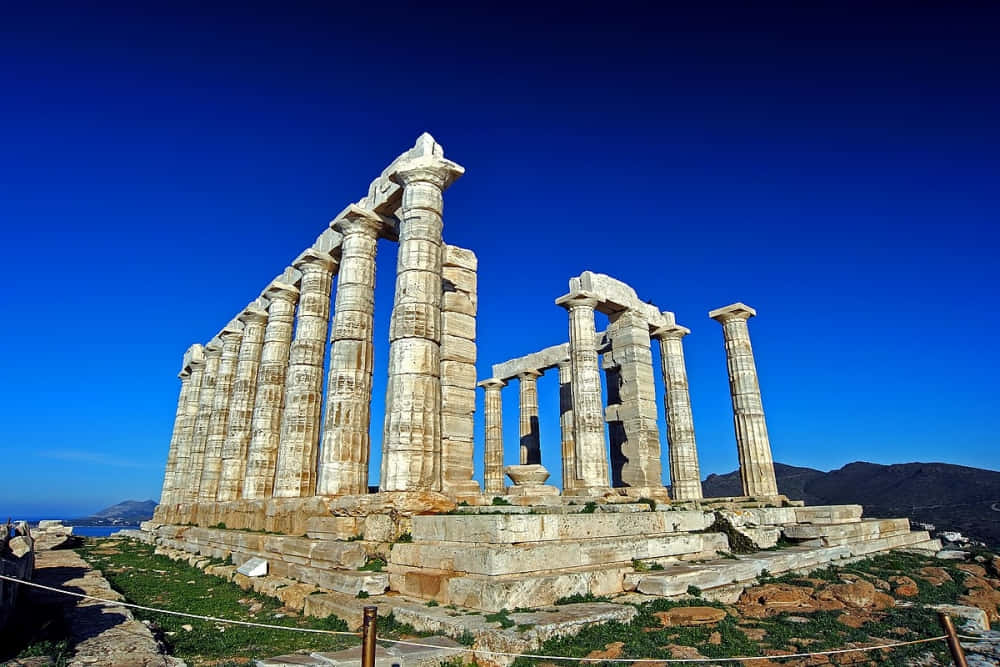 Greek Aesthetic 桌布 壁紙 — 古希臘神廟遺址，高聳的石柱在深藍色的天空下，雄偉地矗立在陽光普照的懸崖邊。