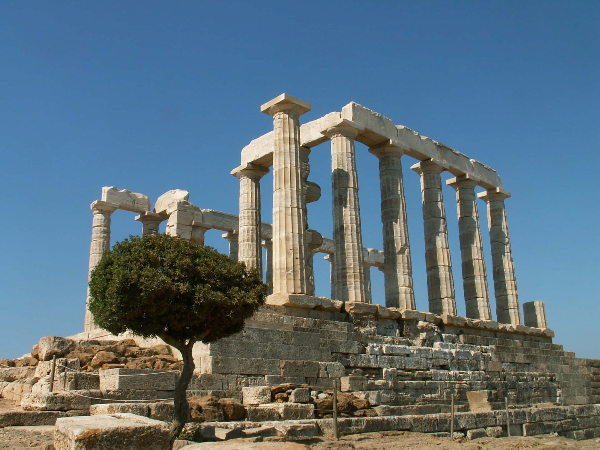 Greece 桌布 壁紙 — 古希臘神廟遺跡，高聳的石柱矗立在清澈的藍天下，由一棵孤獨的綠樹框著。