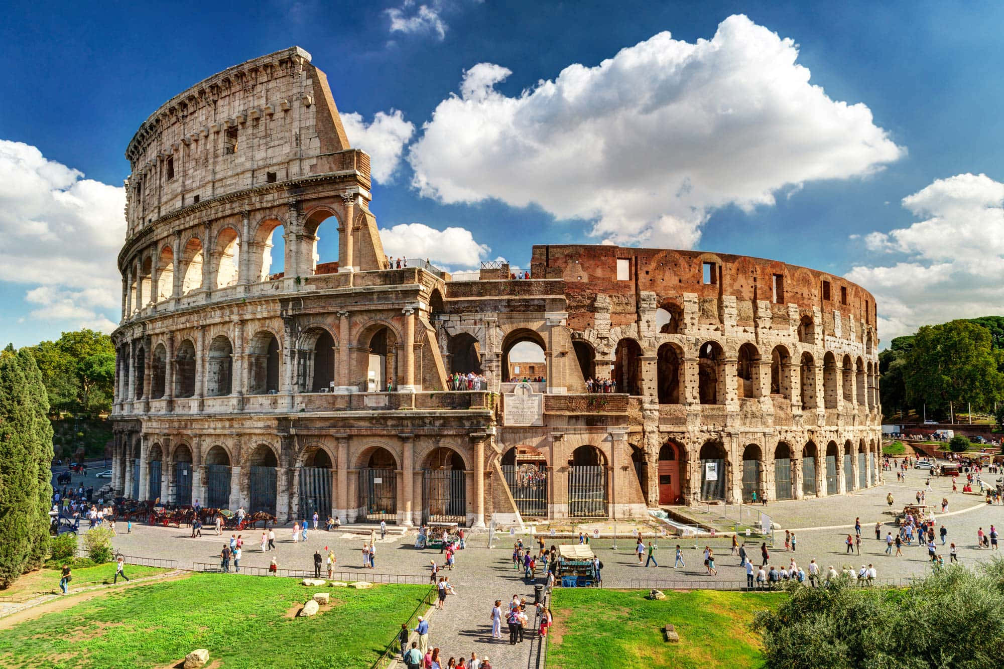 Roman Colosseum 桌布 壁紙 — 古羅馬競技場在明亮的藍天和白雲下雄偉地矗立，遊客們正在探索其歷史遺跡。