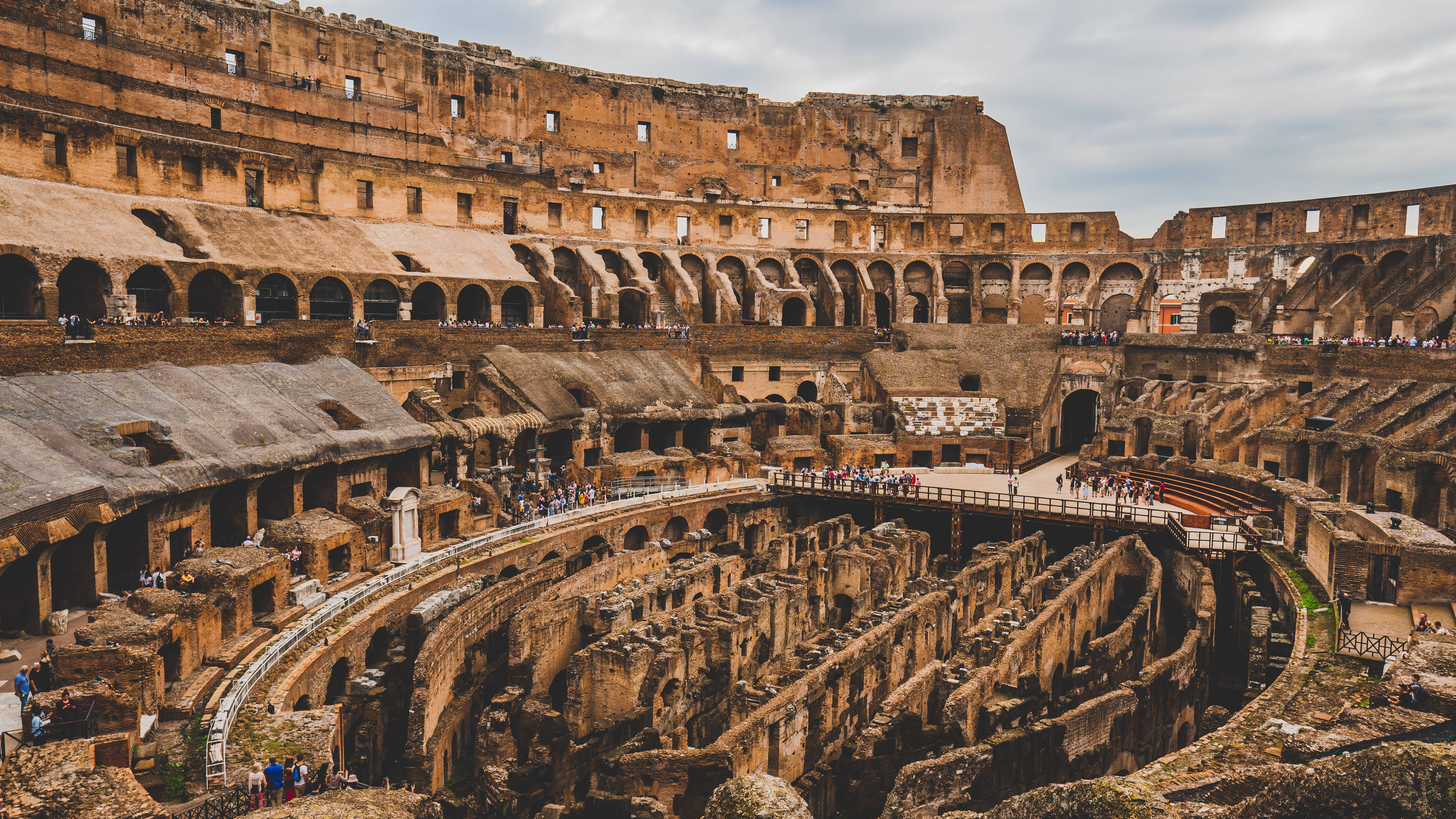 Rome 桌布 壁紙 — 古羅馬競技場遺址，遊客在多雲的天空下探索其廣闊的石頭競技場。