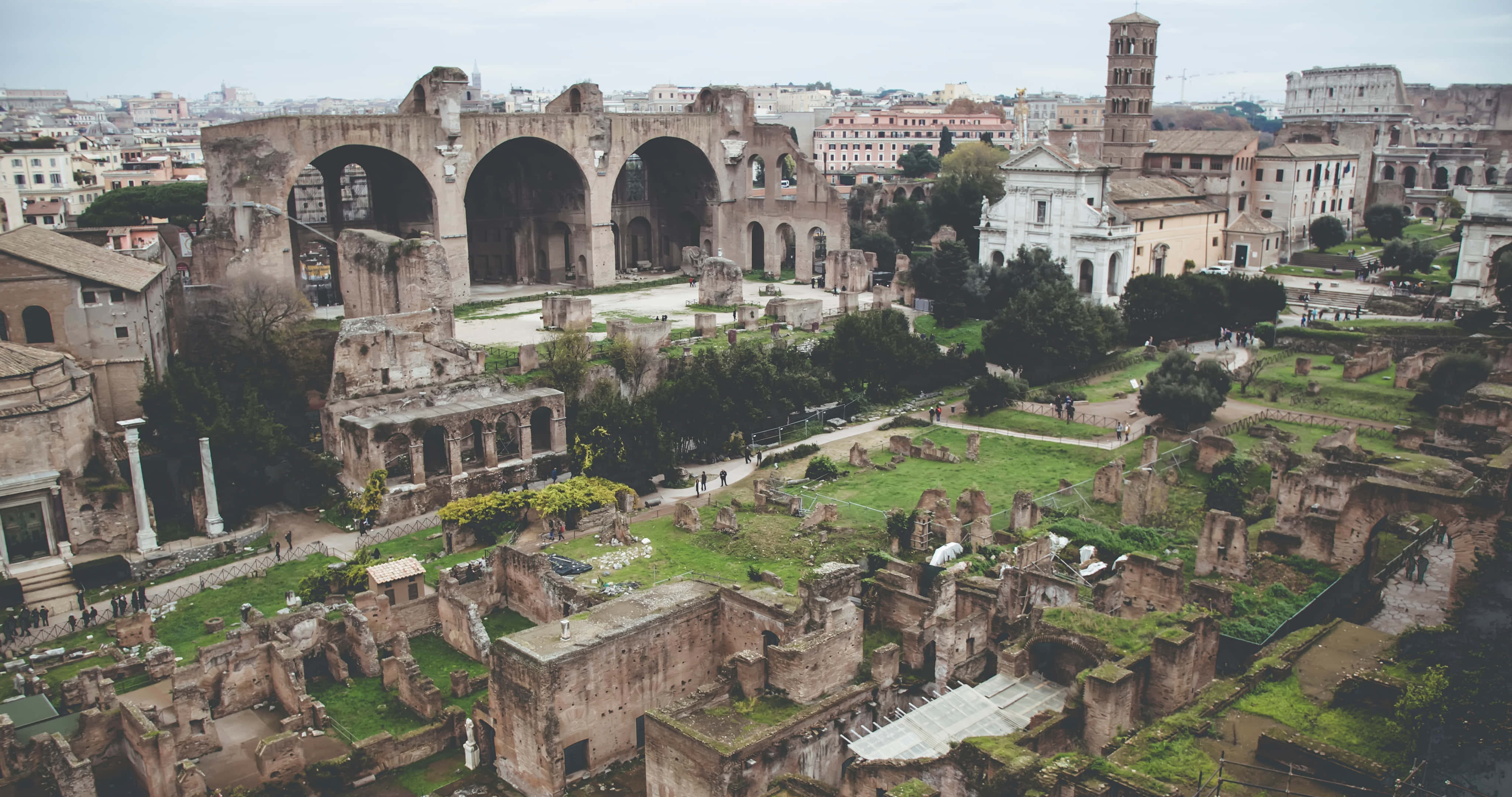 Ancient Roman Desktop 桌布 壁紙 — 古羅馬遺跡，有拱門和圓柱，矗立在綠草和現代建築之間，天空多雲。