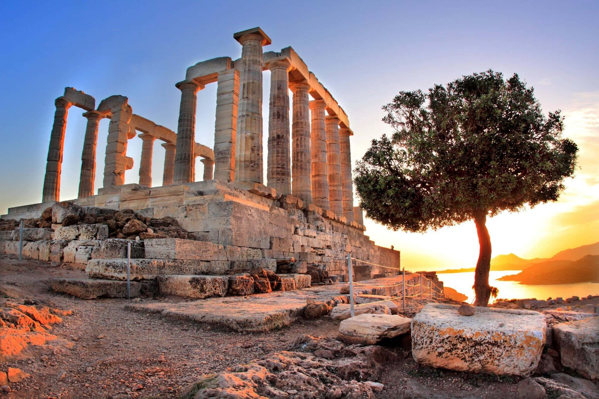 greek 桌布 壁紙 — 古老寺廟遺跡沐浴在金色夕陽光芒中，一條岩石小徑旁有一棵孤樹，俯瞰水面。