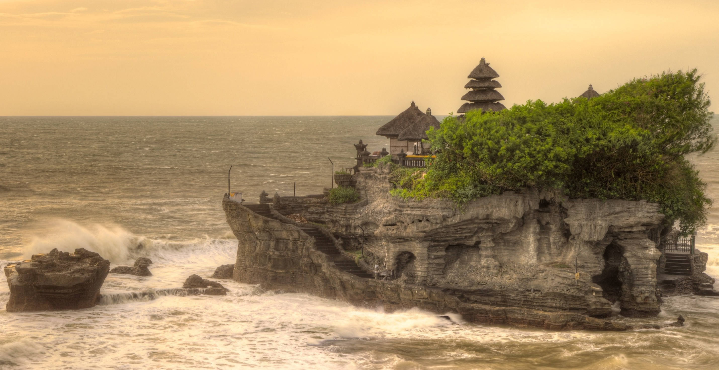 印尼 桌布 壁紙 — 古老峇里寺廟矗立於岩石島上，海浪在金色夕陽天空下拍打其基部。