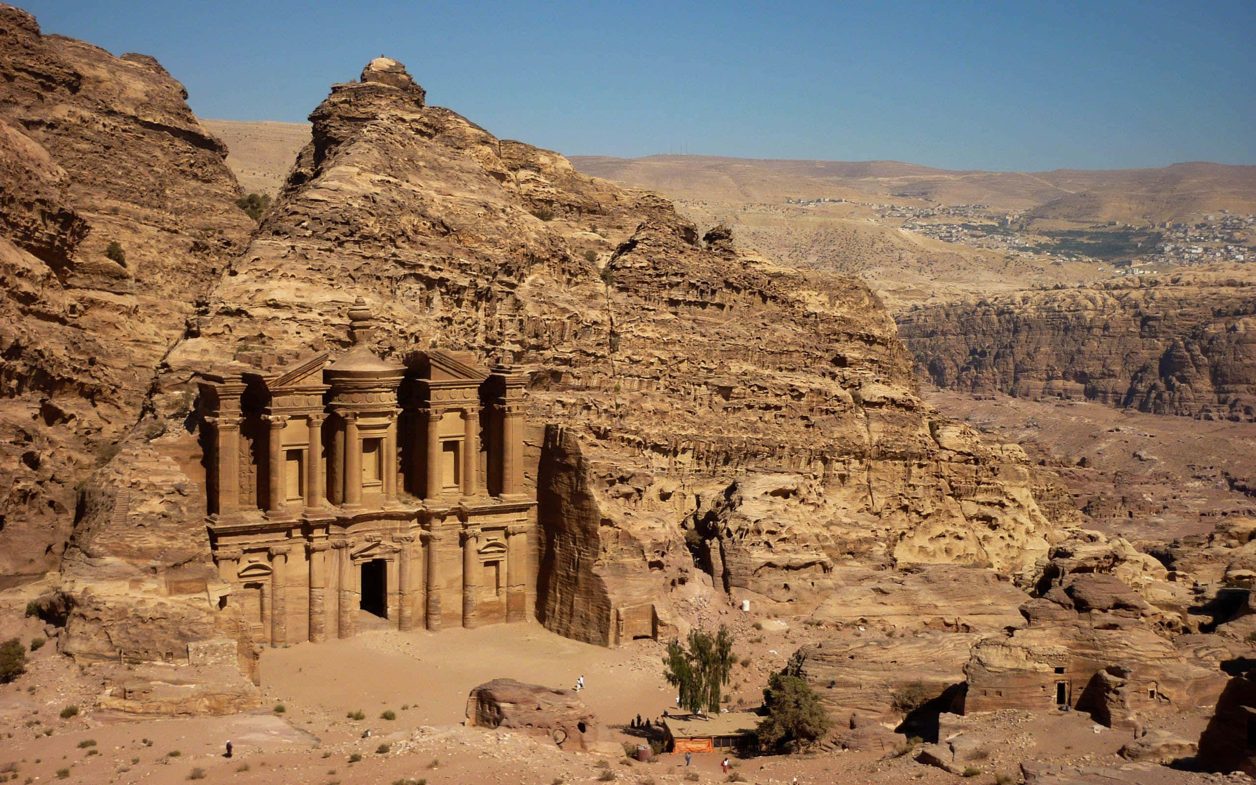 Jordan 桌布 壁紙 — 古老玫瑰砂岩寺廟雕刻在沙漠懸崖上，藍天白雲，遠處有山谷小鎮。