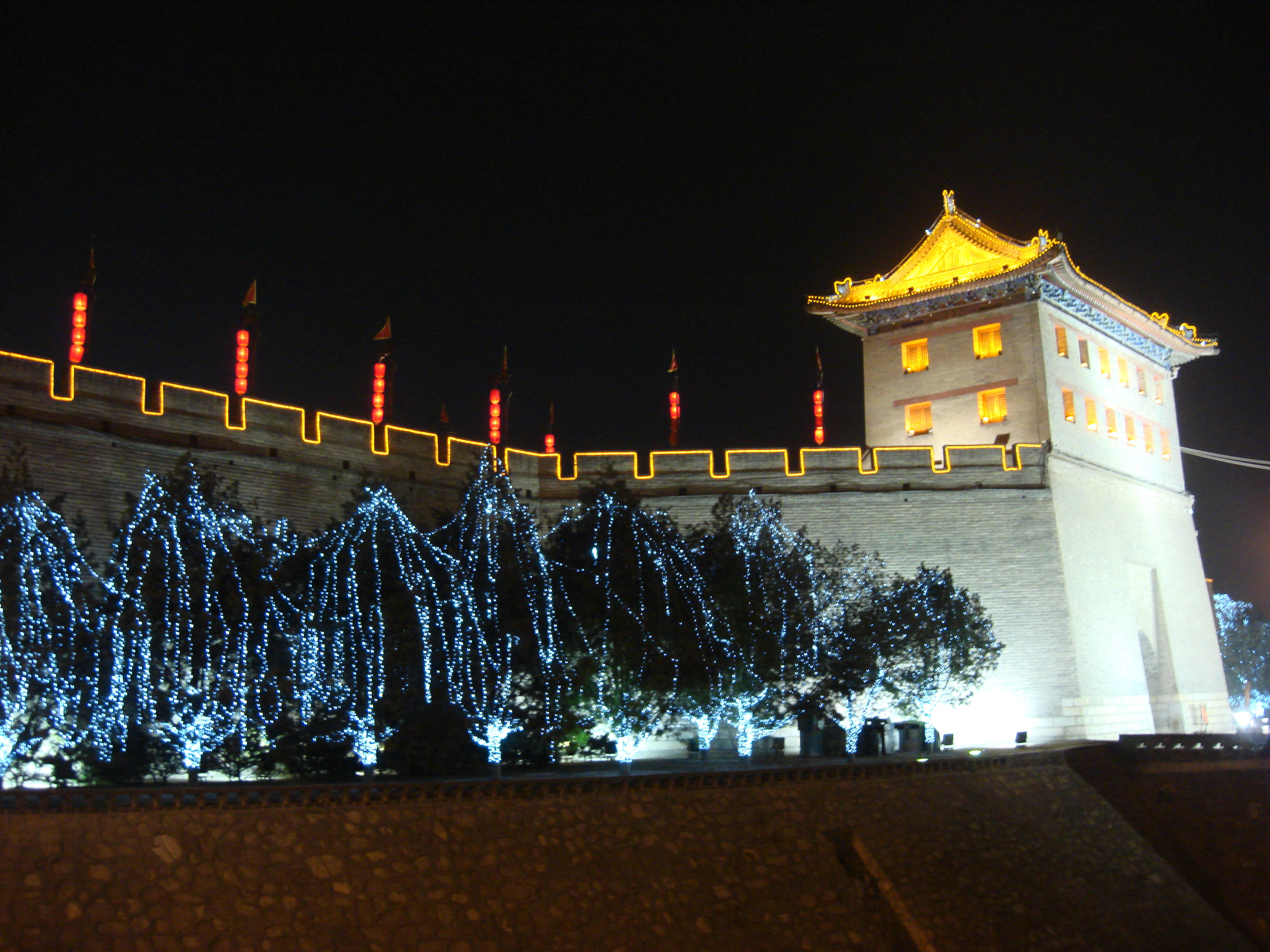 Xian 桌布 壁紙 — 古老的中國城牆在夜晚被照亮，塔樓上閃爍著金色燈光，下方的樹木上掛著藍色串燈。
