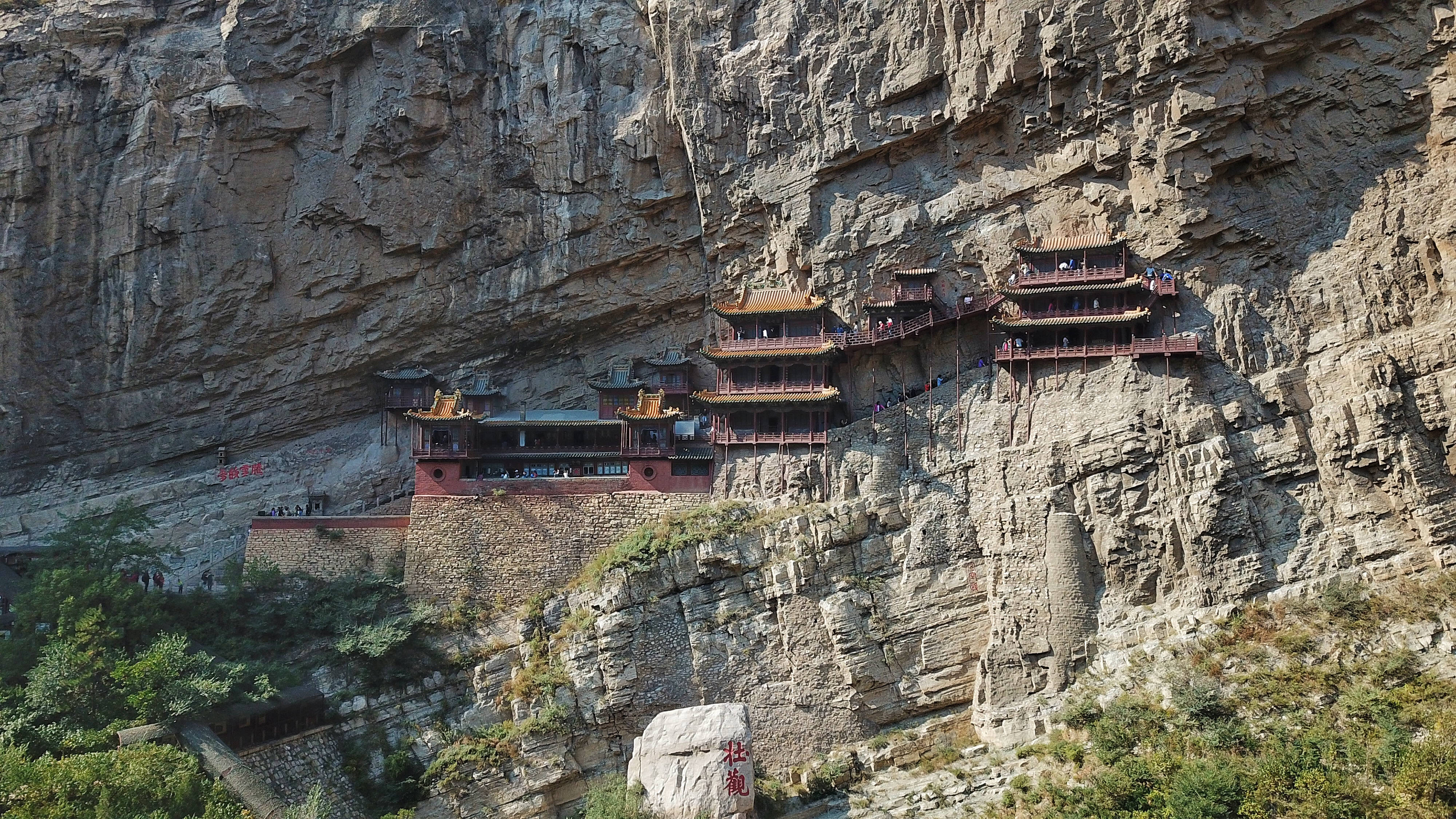 中國 桌布 壁紙 — 古老的中國寺廟建築群緊貼著陡峭的懸崖面，有木製走道和紅色屋頂，襯托著崎嶇的石頭。