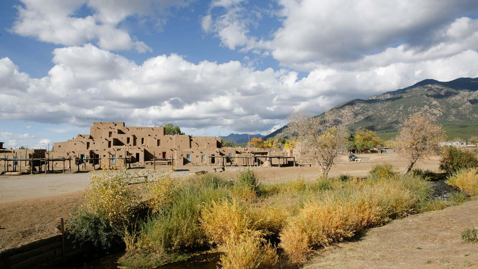 New Mexico 桌布 壁紙 — 古老的土坯普韋布洛遺址矗立在明亮的藍天下，有蓬鬆的白雲和遠山。