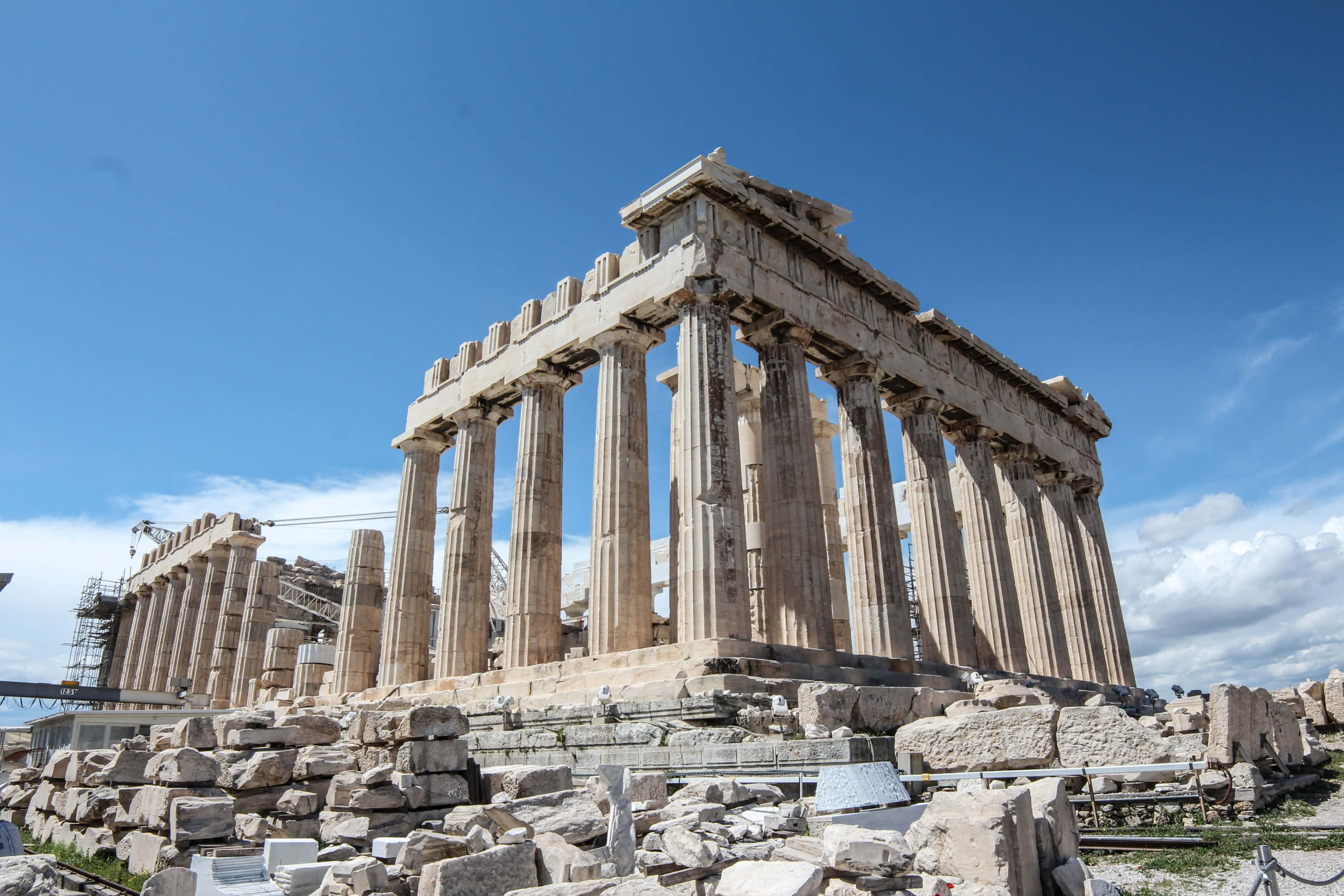 greek 桌布 壁紙 — 古老的帕德嫩神廟在清澈的藍天下，伴隨著散落的雲朵和周圍的石頭廢墟，雄偉地矗立著。
