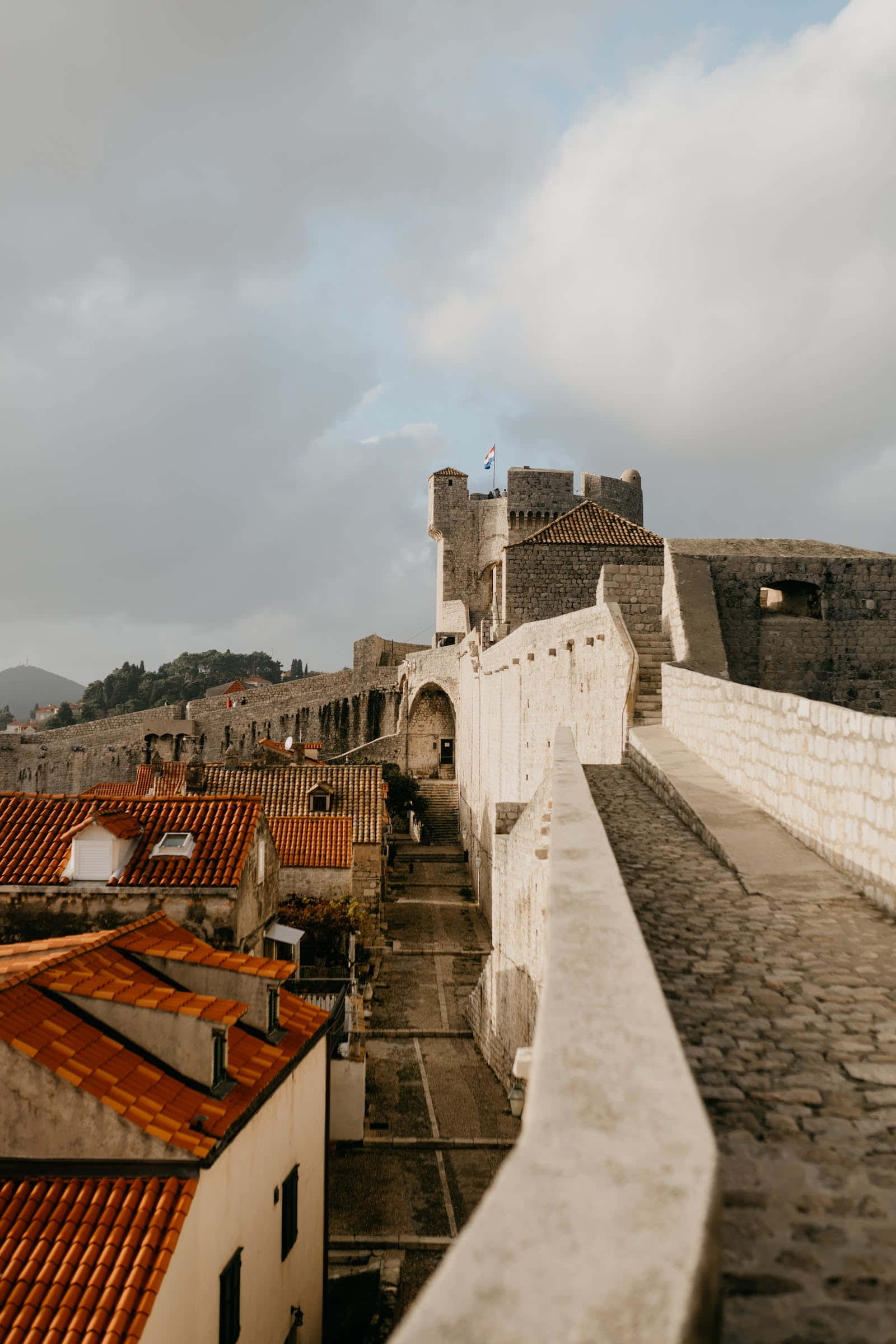 Middle Ages 桌布 壁紙 — 古老的石堡壘牆俯瞰著一條狹窄的鵝卵石街道，兩旁是鋪著陶土瓦頂的建築物，天空多雲。