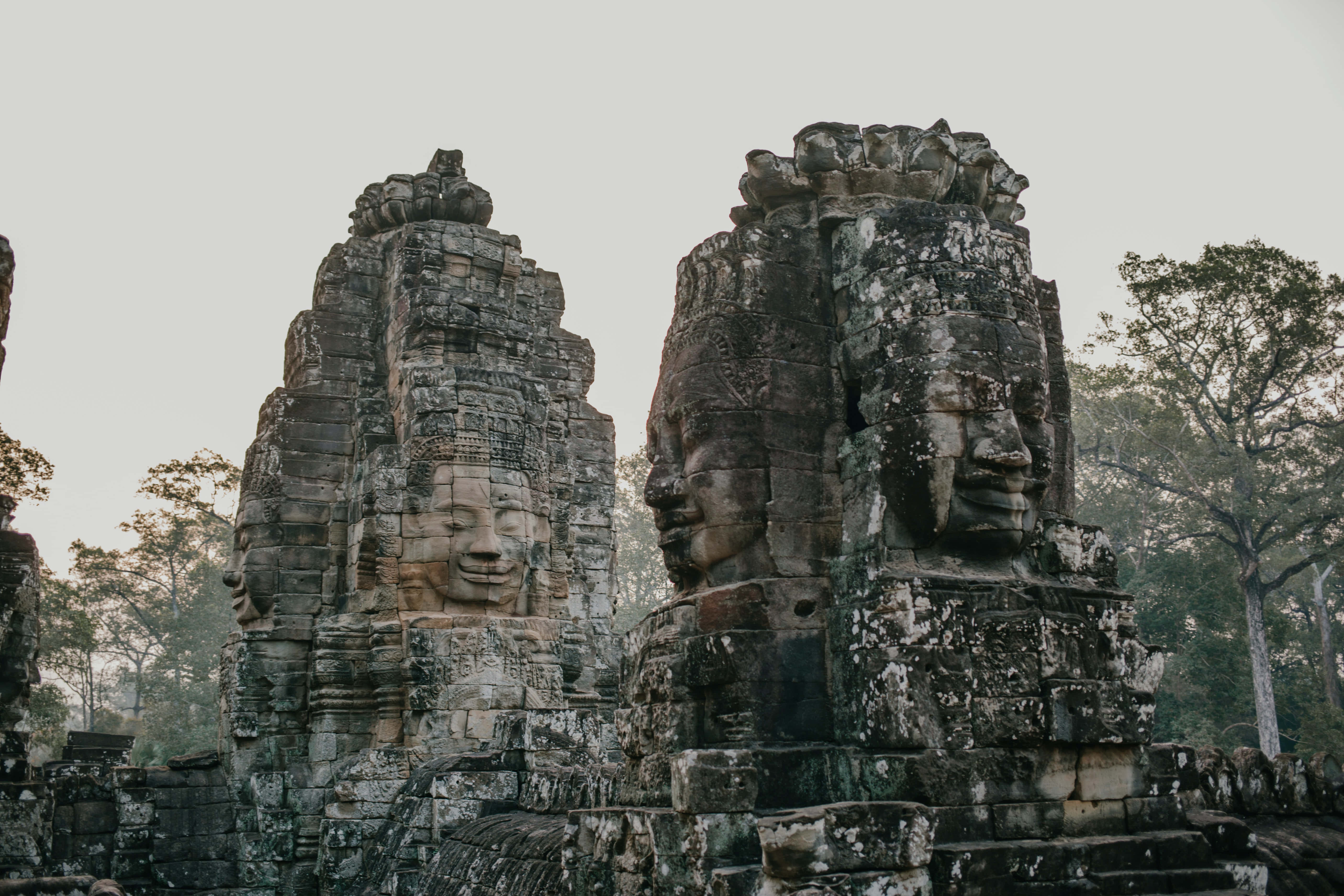 Cambodia 桌布 壁紙 — 古老的石塔上雕刻著寧靜的面孔，在柔和的陰天下從叢林樹葉中升起。