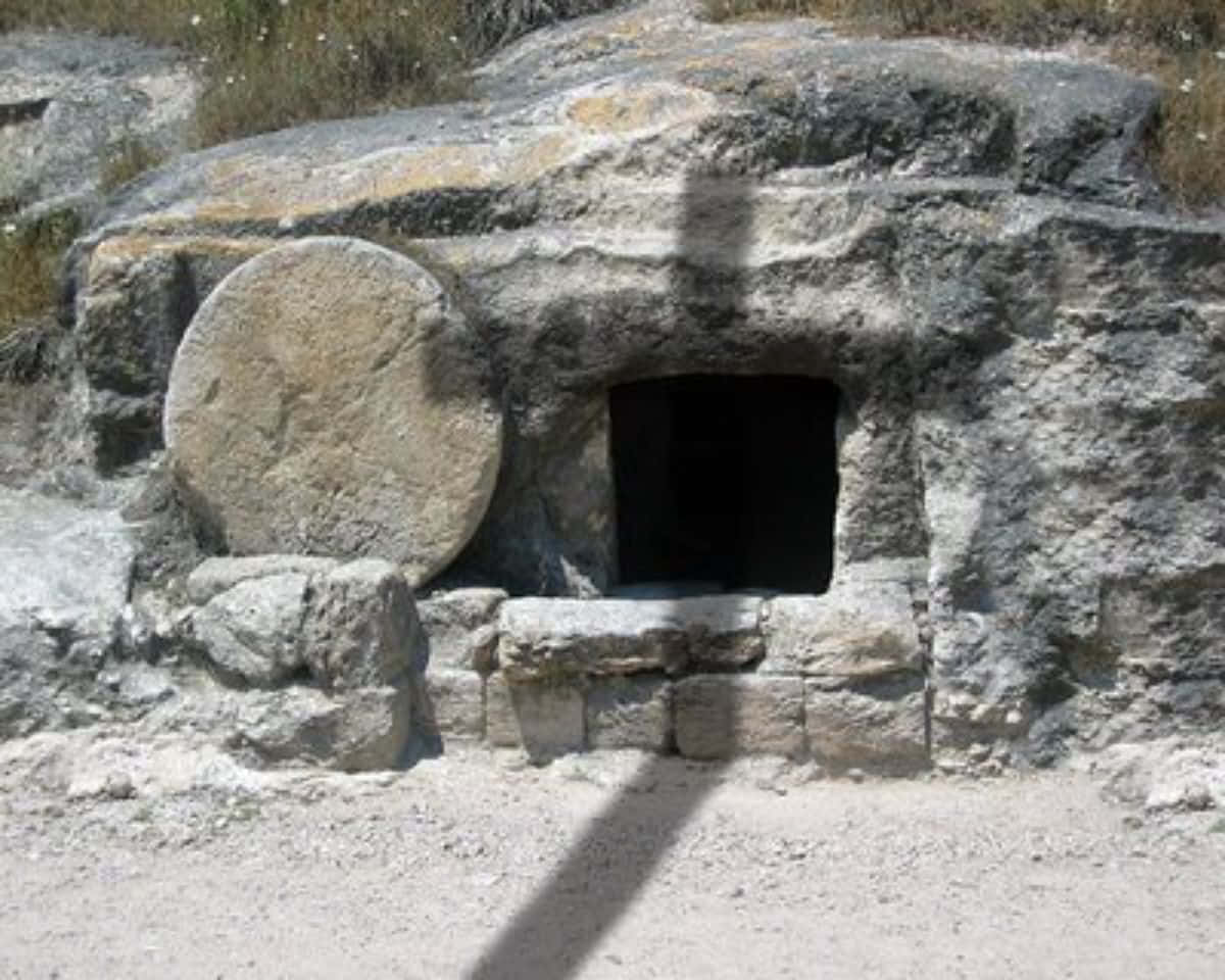 Empty Tomb 桌布 壁紙 — 古老的石墓，帶有大型圓形蓋子和黑暗入口，雕刻在陽光普照的岩石地形中。